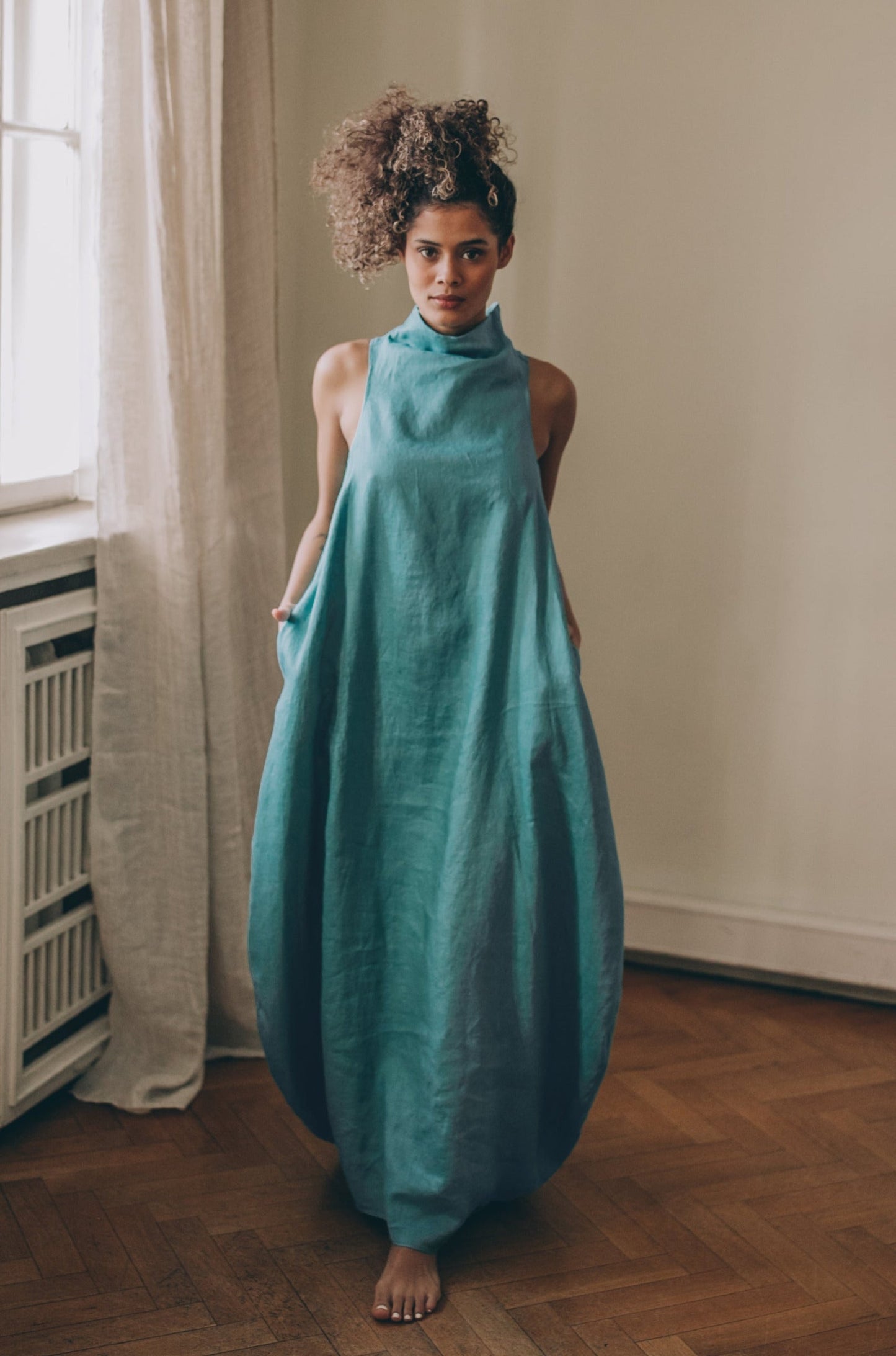 Woman wearing blue green sleeveless cowl neck linen dress with asymmetric front and side pockets indoors