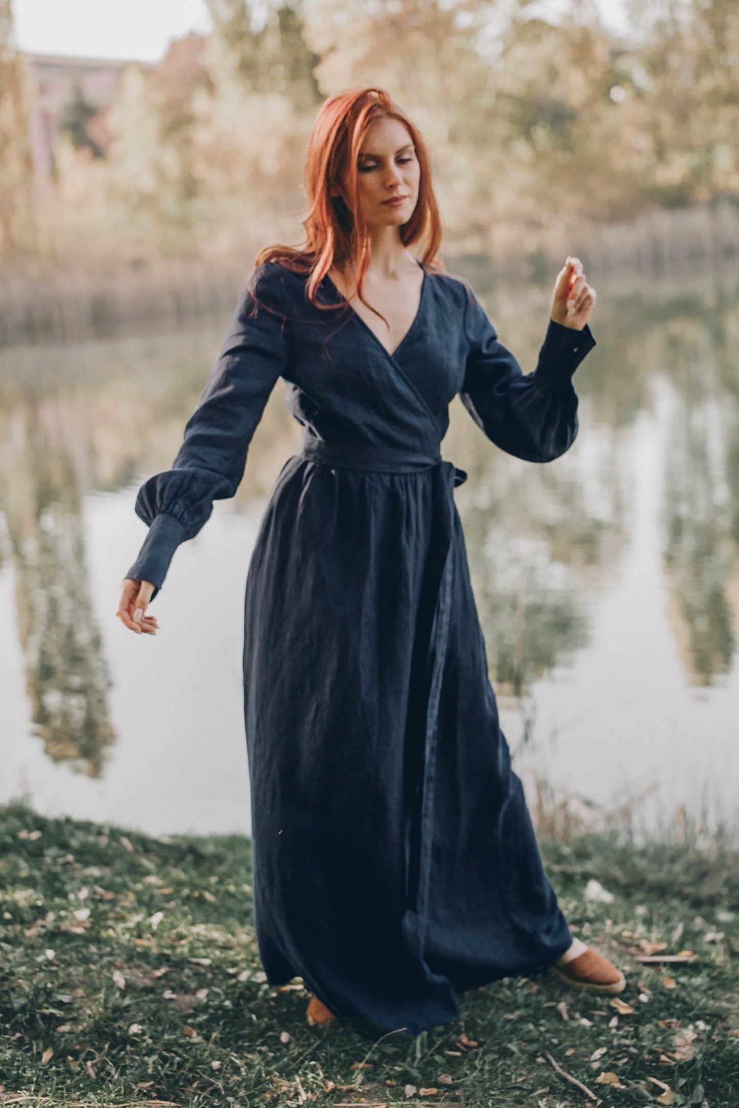 Woman wearing navy blue heavyweight wrap linen dress with tie waist and long cuff sleeves outdoors by a lake