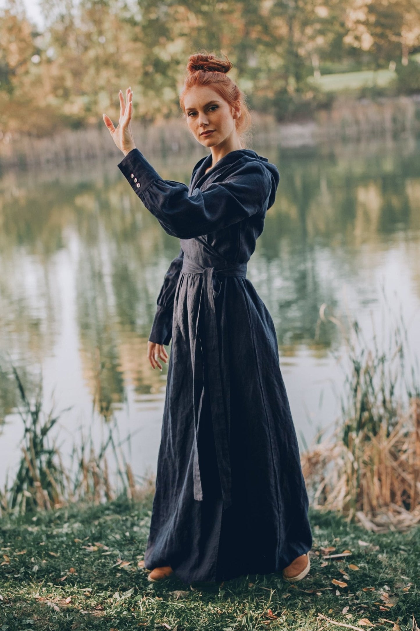 Woman wearing night blue maxi linen wrap dress with hood standing outdoors by a lake