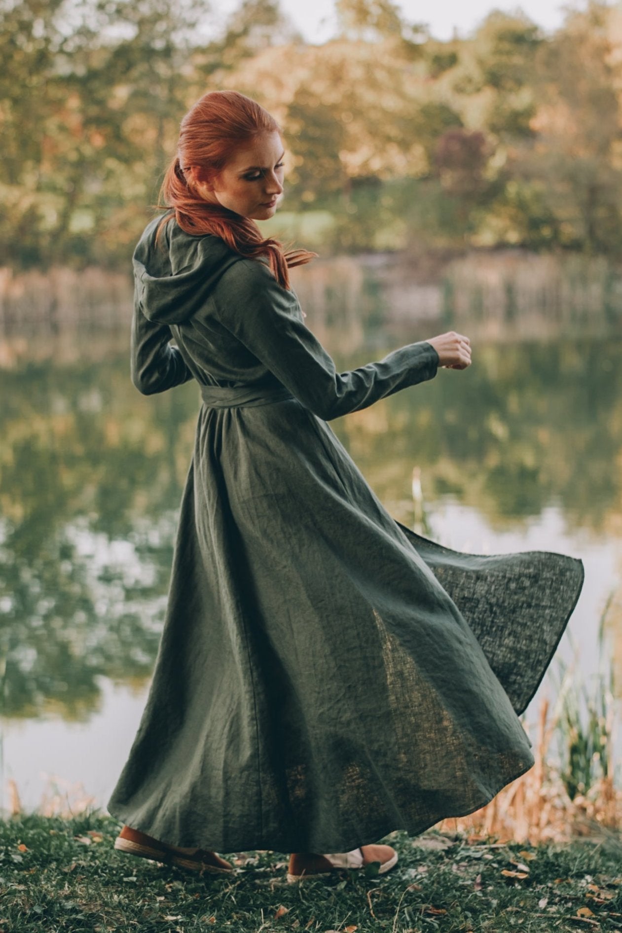 Woman wearing a green linen A-line midi dress with hood standing by a lake