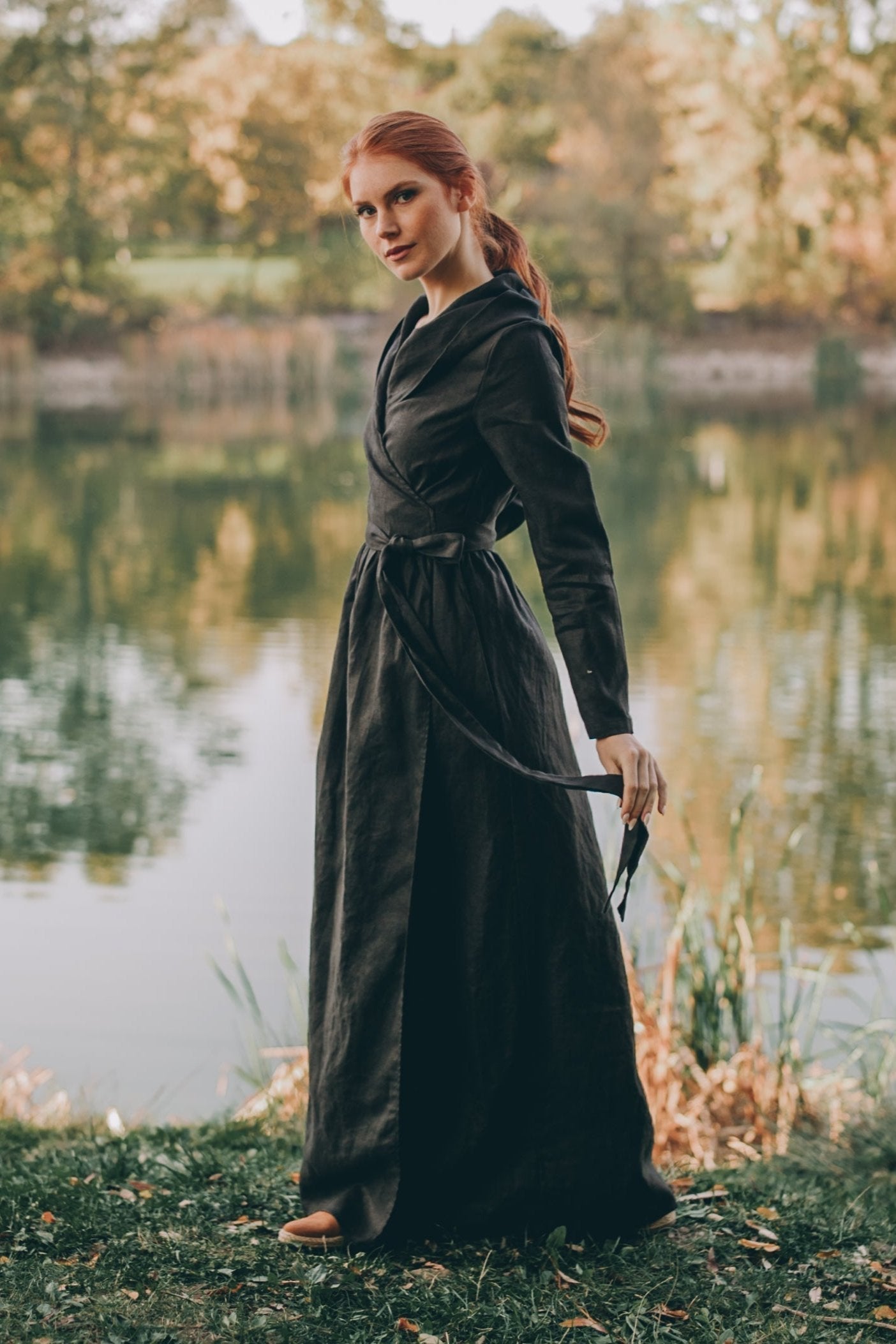 Woman wearing a long sleeve linen wrap dress with hood standing outdoors by a lake