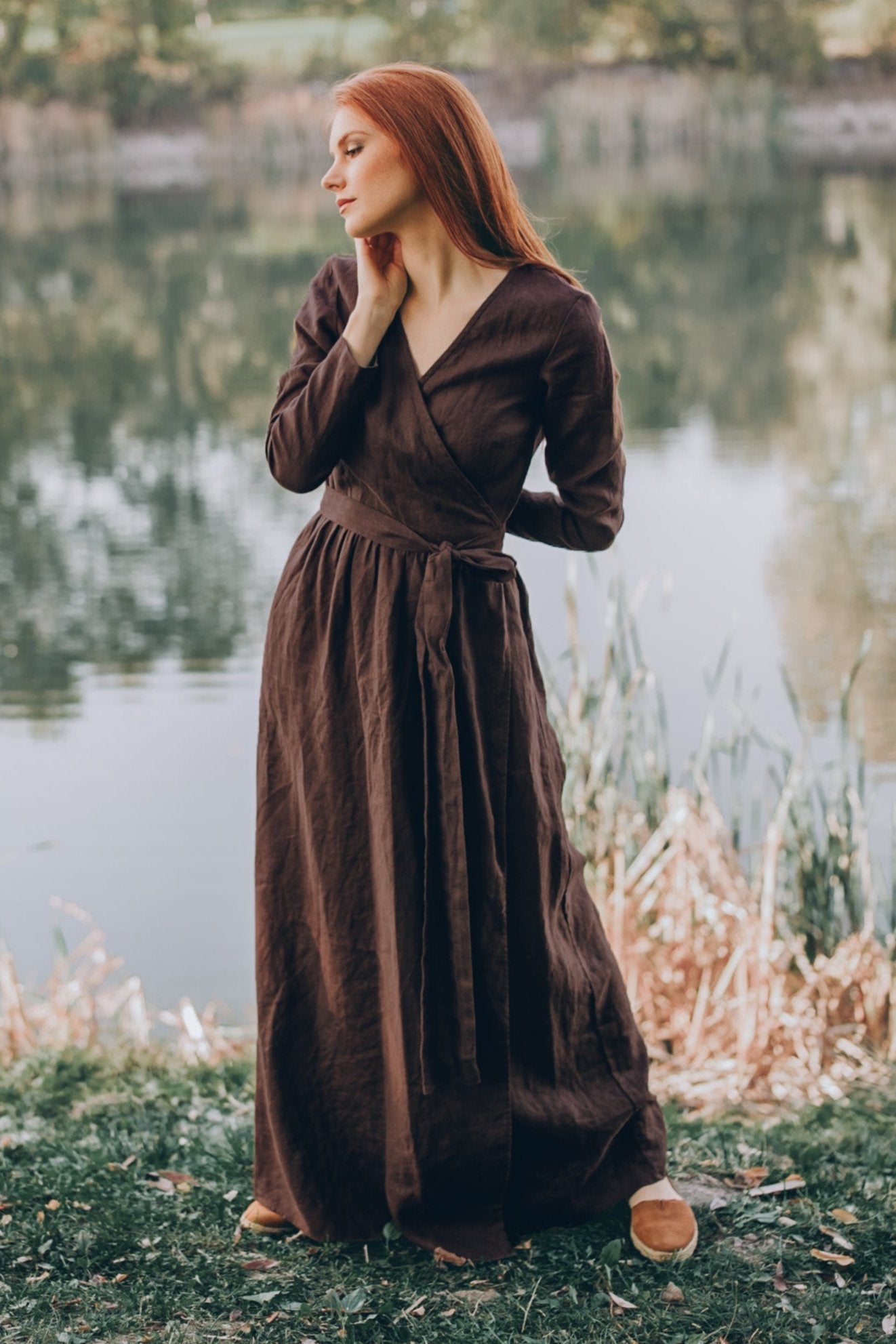 Woman wearing brown long sleeve linen wrap dress standing near water outdoors