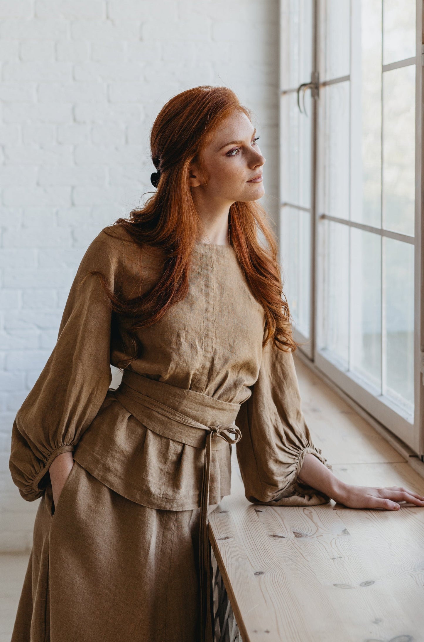 Woman wearing brown puff sleeve linen top with belt standing by window