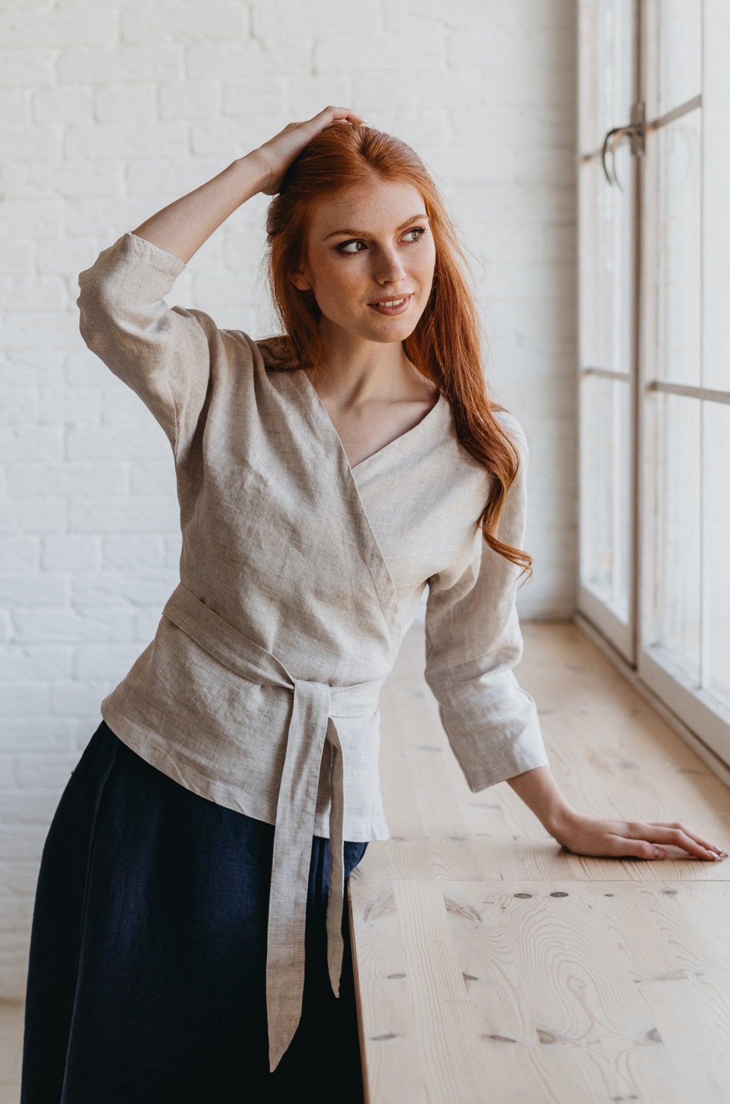 Woman wearing reversible minimalist linen wrap top with tie waist and elbow-length sleeves standing by window