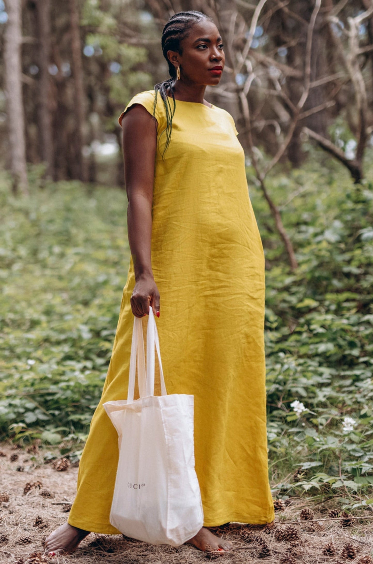 Woman wearing mustard long linen dress with cap sleeves standing in forest holding white tote bag