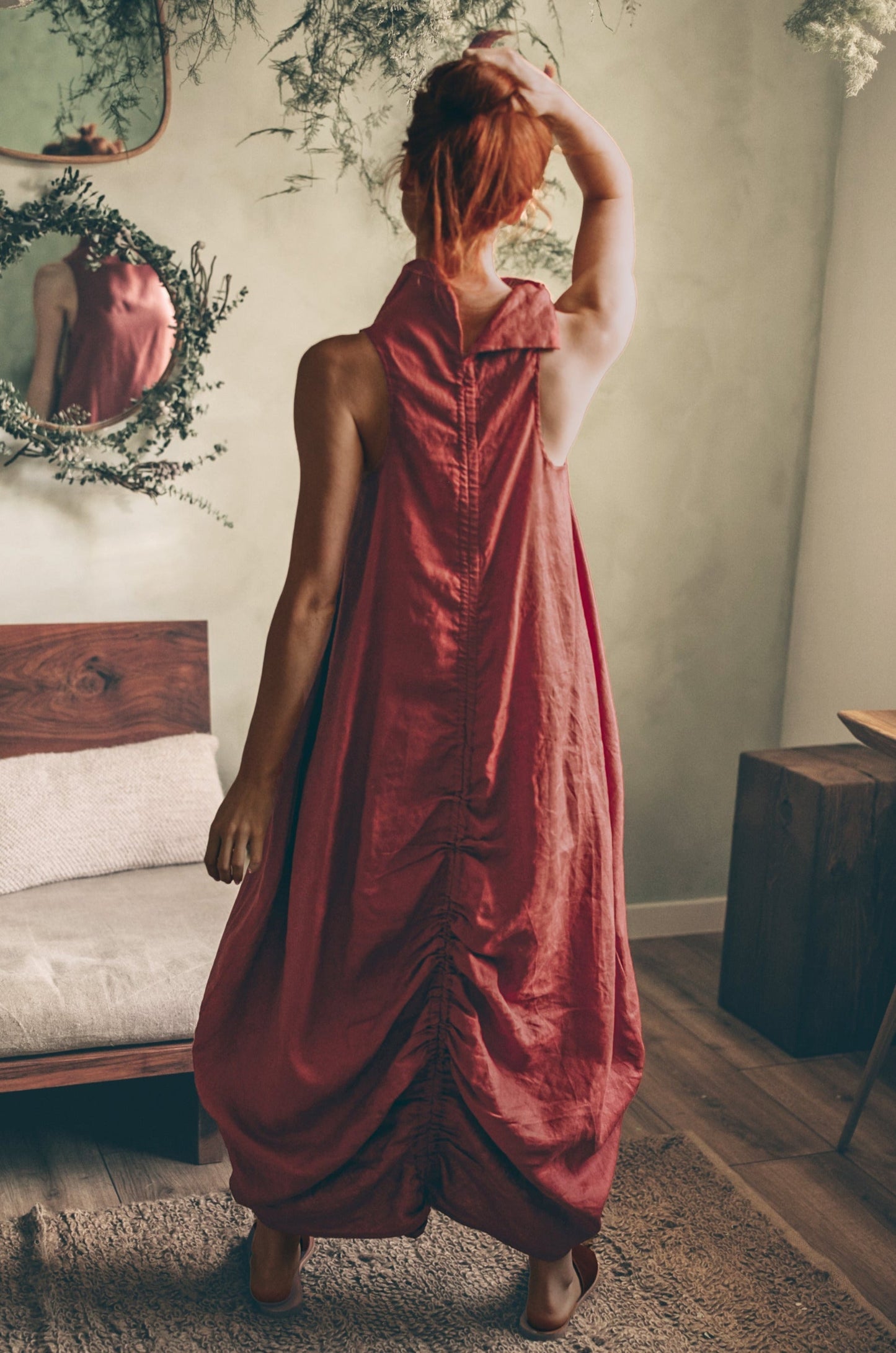 Back view of a Moroccan Red sleeveless cowl neck linen dress with ruched adjustable back and side pockets worn by a woman indoors