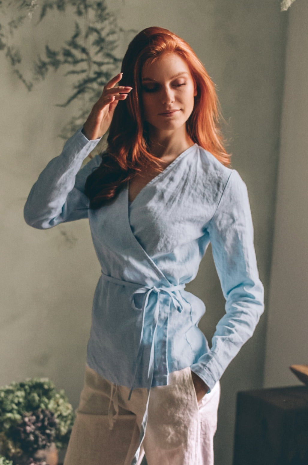 Woman wearing light blue linen wrap shirt with tie belt and full-length sleeves indoors