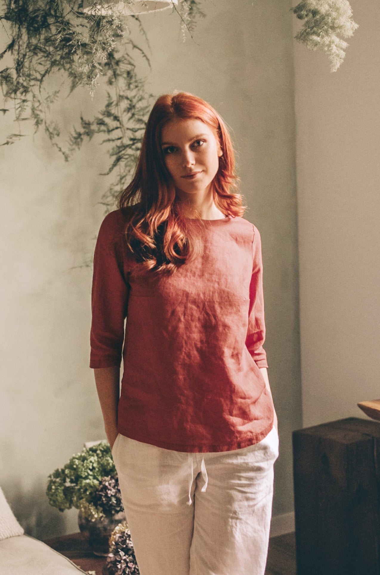 Woman wearing rust-colored essential linen top with 3/4 sleeves and white linen pants in natural light setting