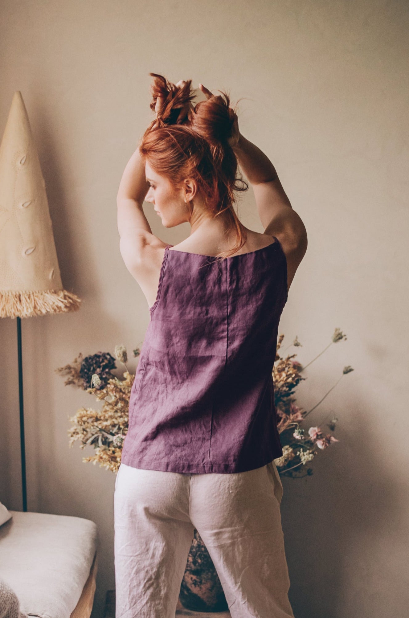 Woman wearing purple straight neck linen top with delicate straps and white linen pants standing indoors with hands raised