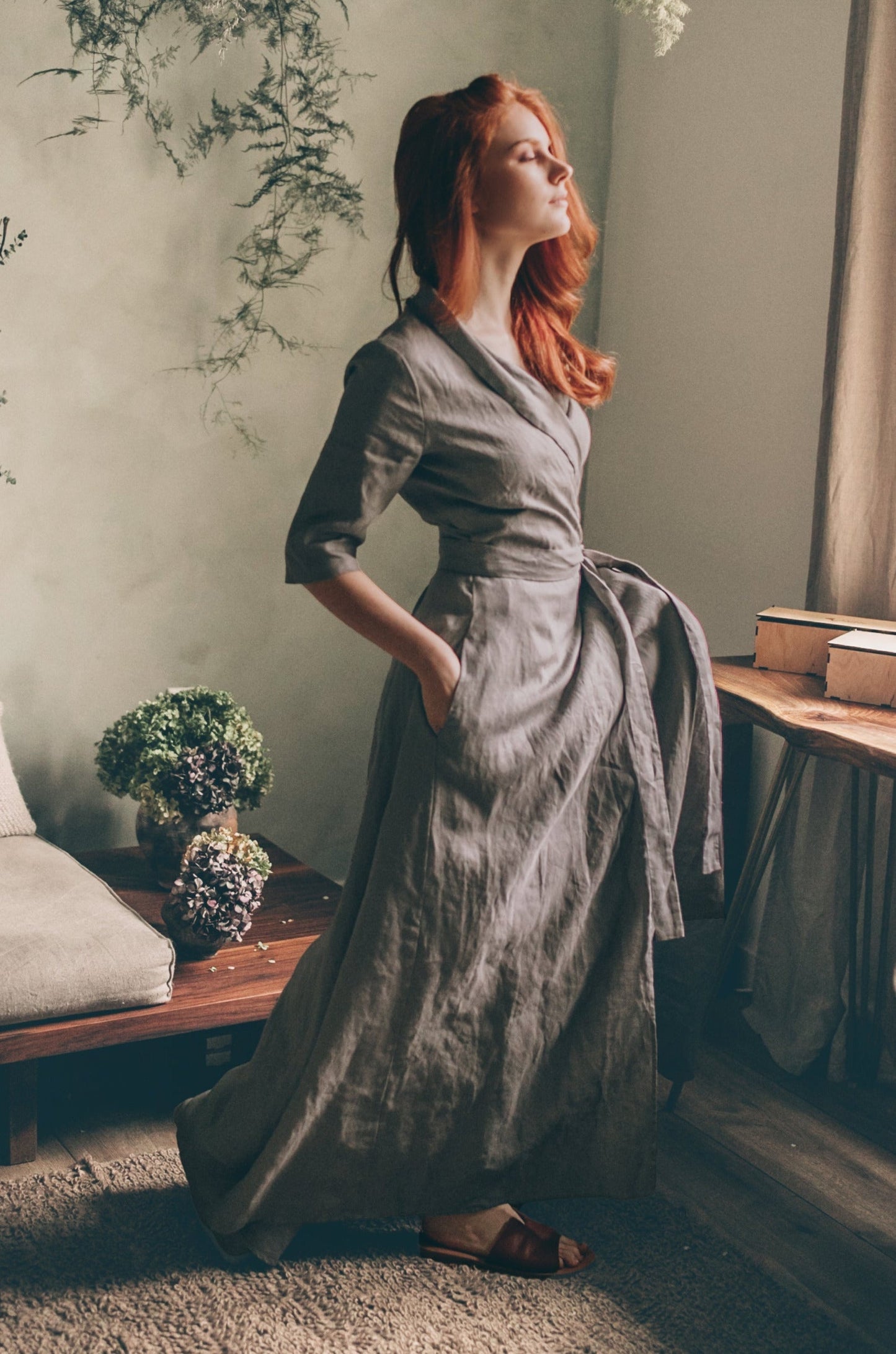 Woman wearing a grey linen A-line dress with tie waist standing indoors by window