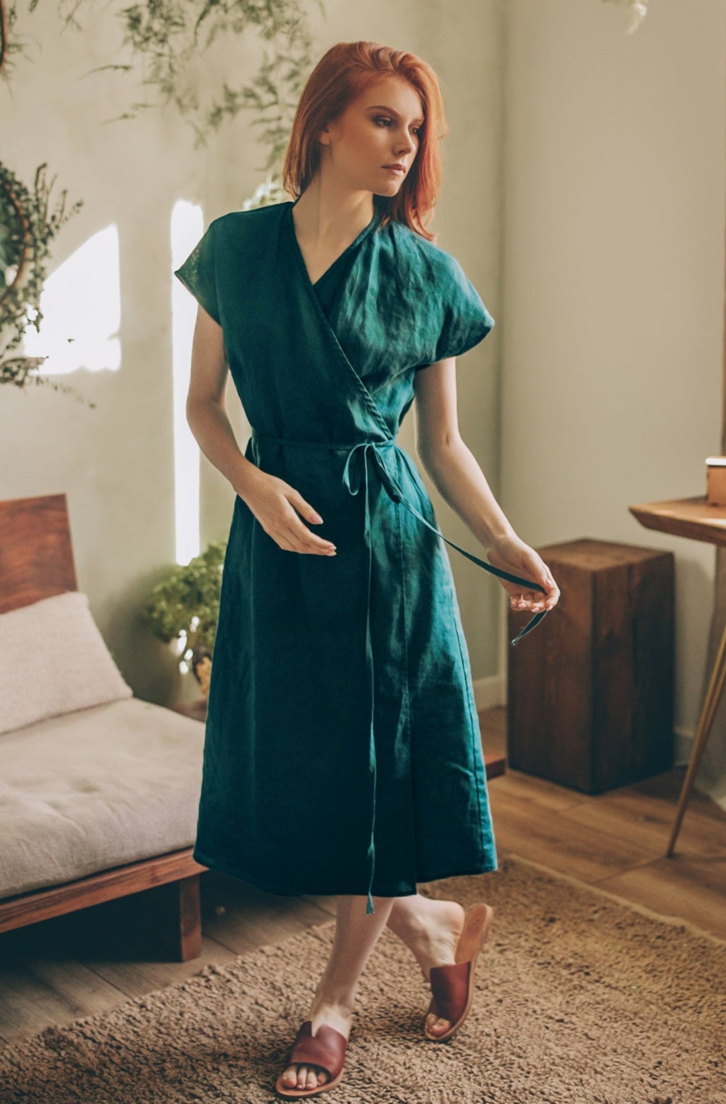 Woman wearing green cap sleeve linen wrap dress with tie waist in cozy room