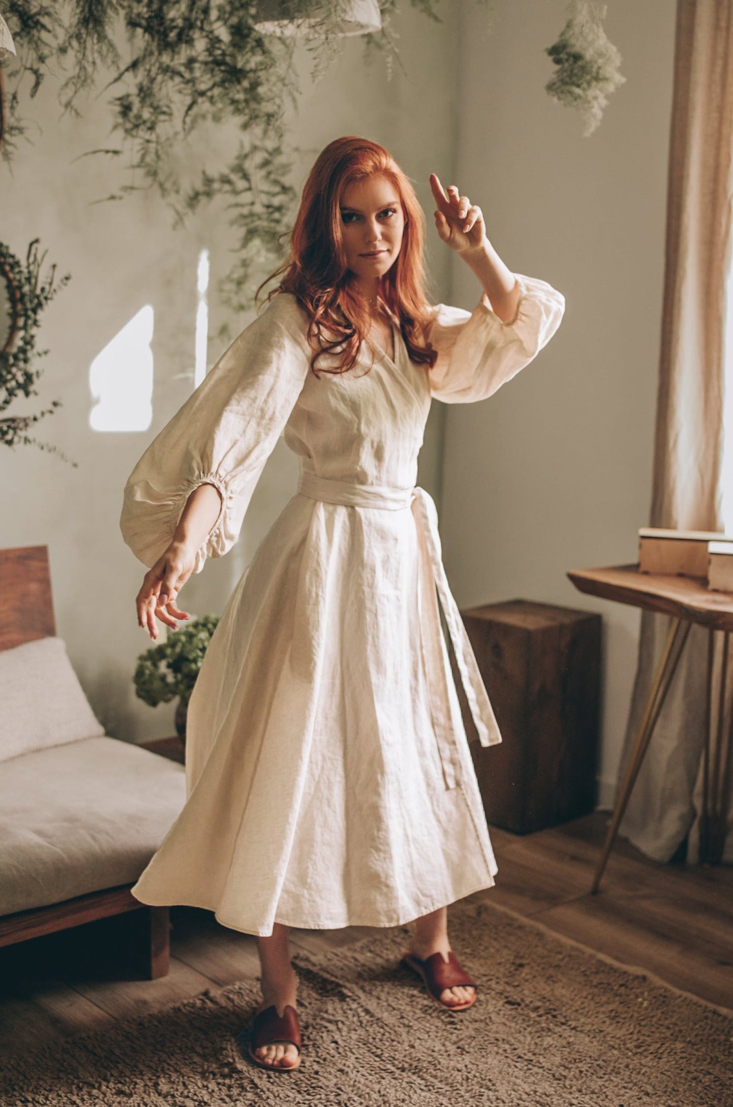 Woman wearing cream linen wrap dress with puff sleeves and tie belt in a cozy room