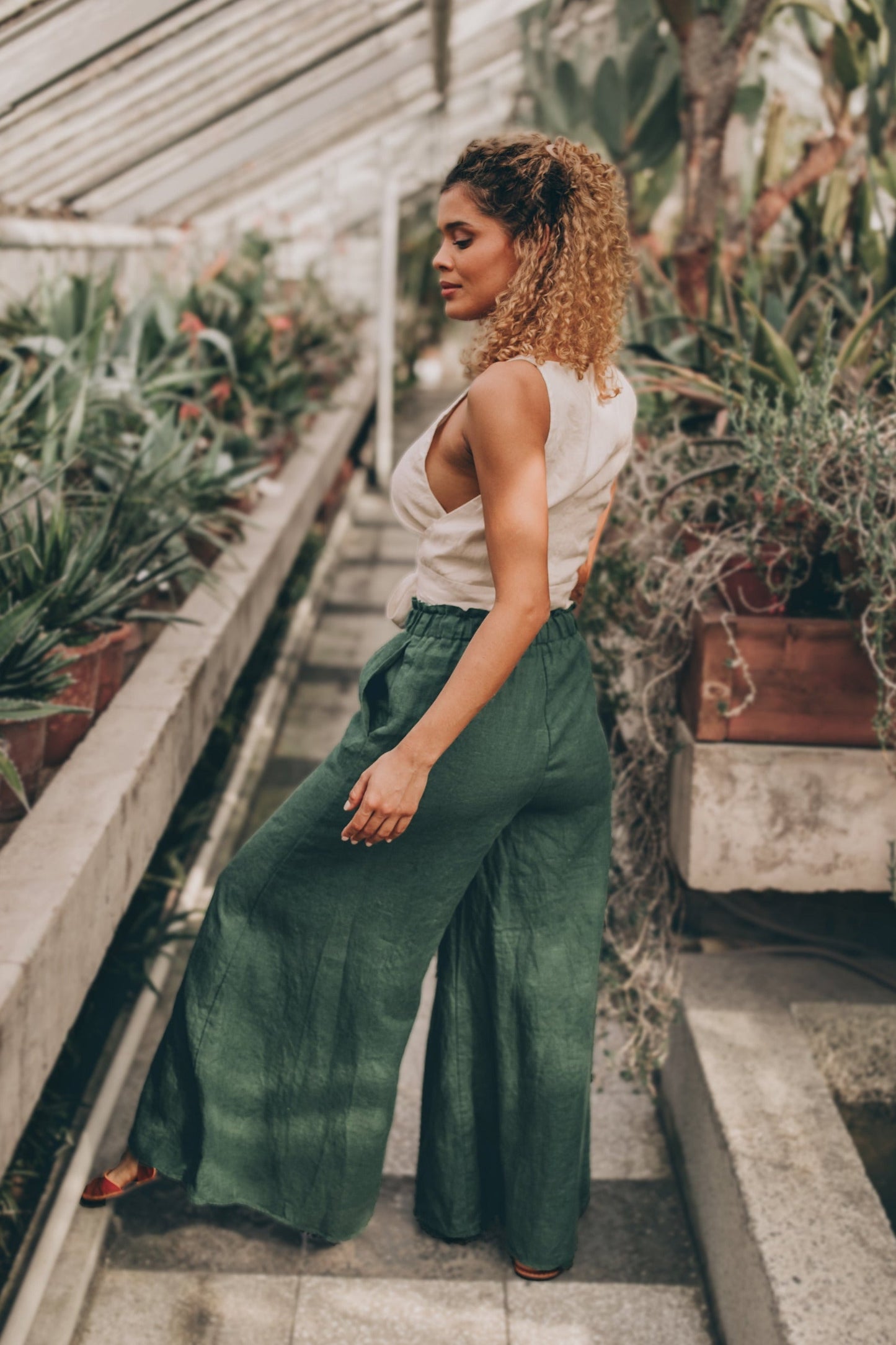 Woman wearing heavyweight linen wide leg green pants with elastic waistband and pockets in greenhouse setting