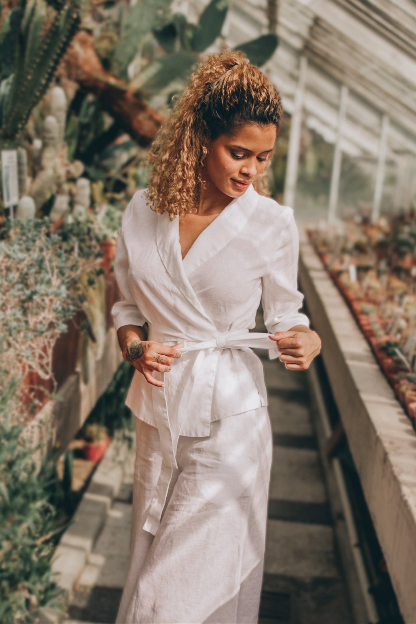 Woman wearing white linen wrap top with shawl neck and tie belt in a greenhouse setting
