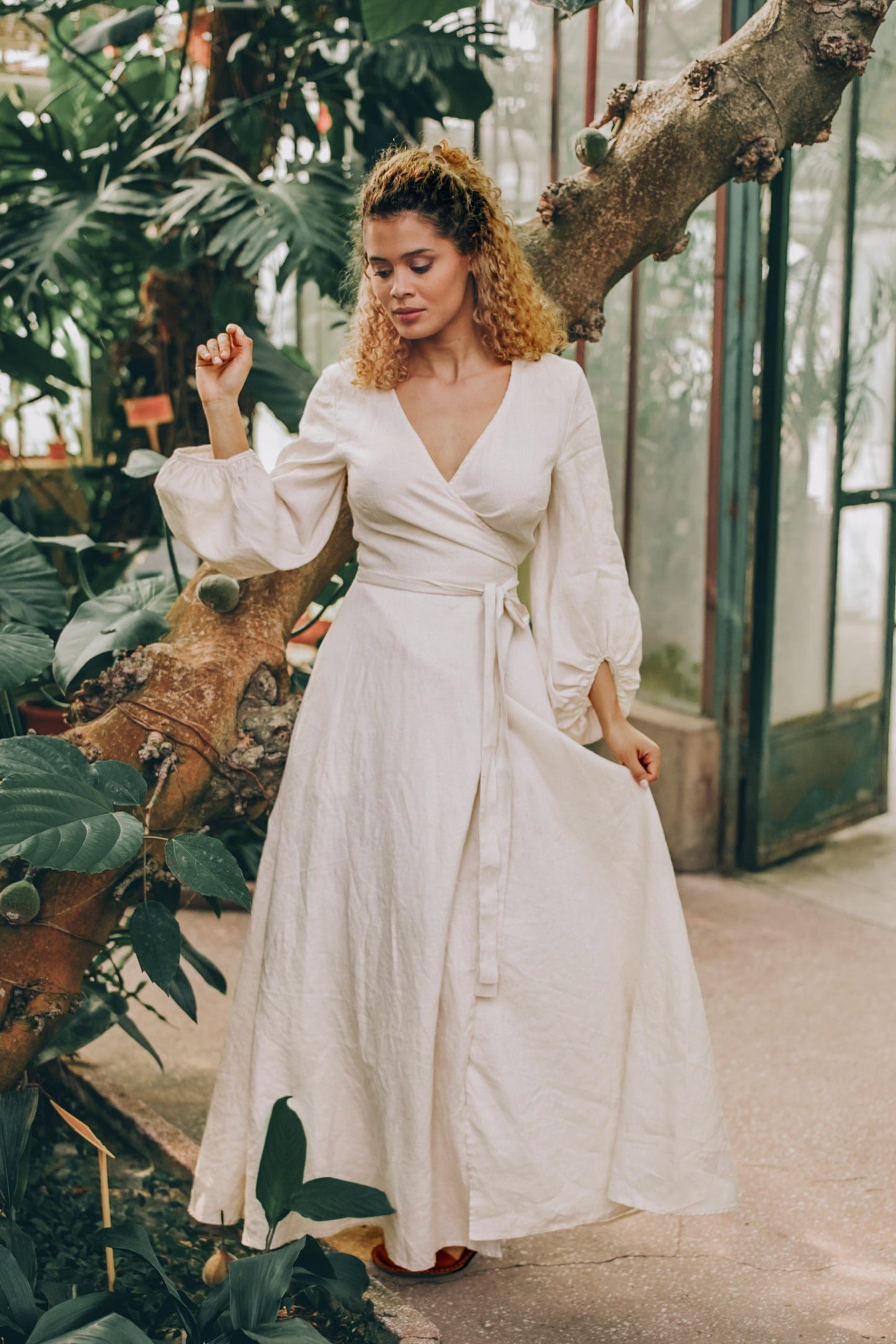 Woman wearing cream linen maxi dress with puff sleeves and tie belt in indoor garden setting