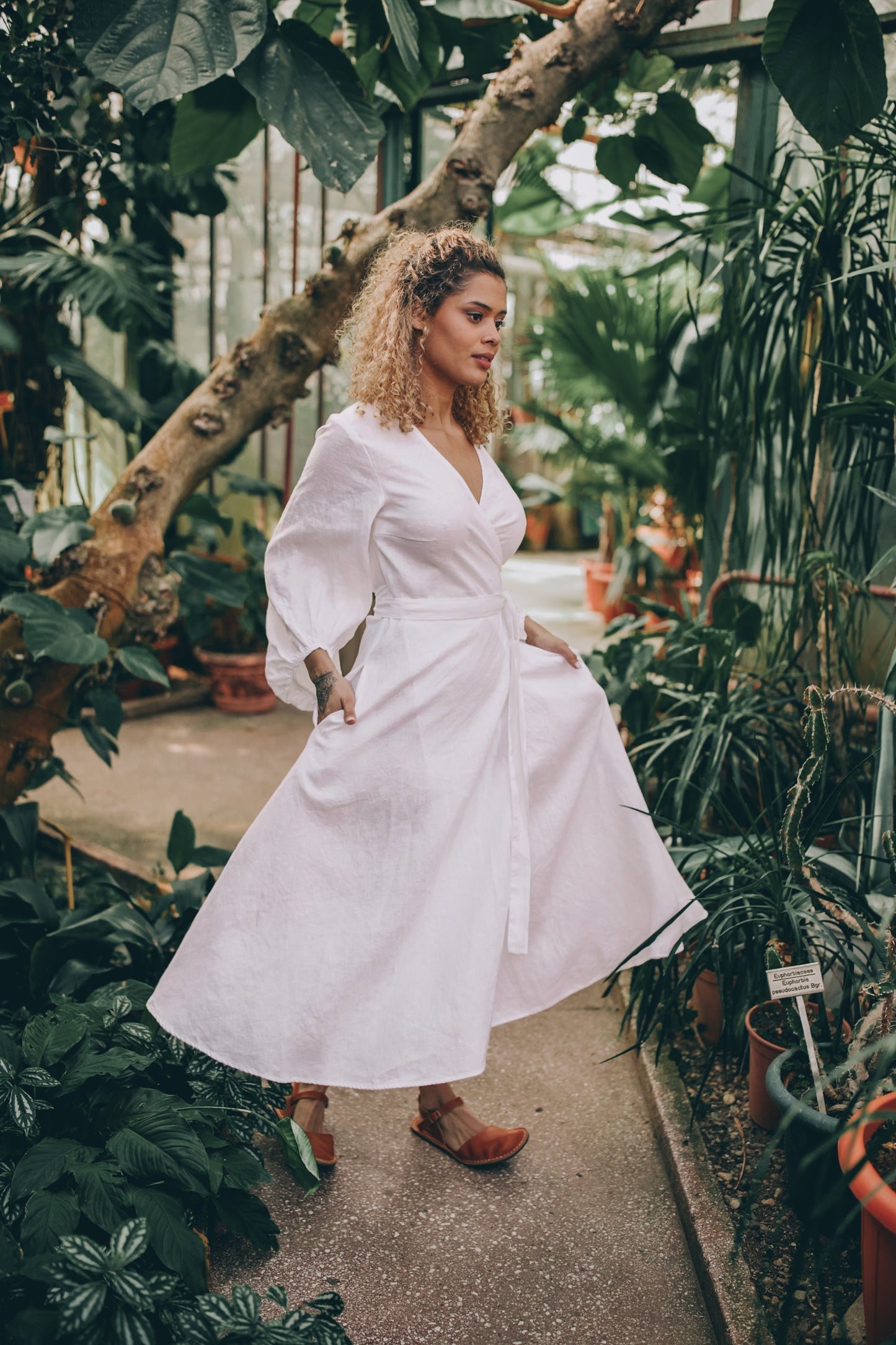 Woman wearing white linen wrap dress with puff sleeves and tie belt in indoor garden