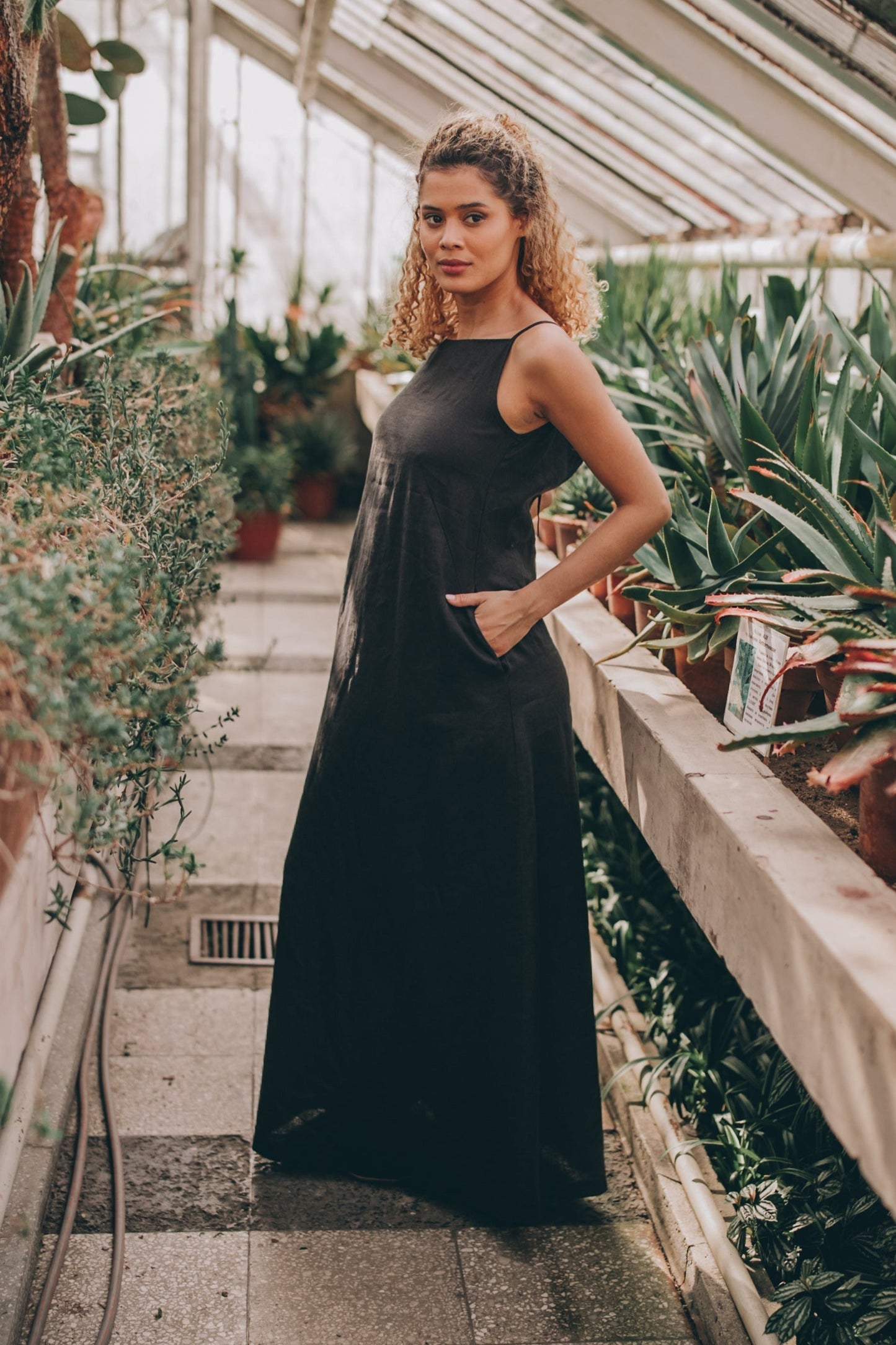 Woman wearing black sleeveless linen maxi dress with pockets in greenhouse setting