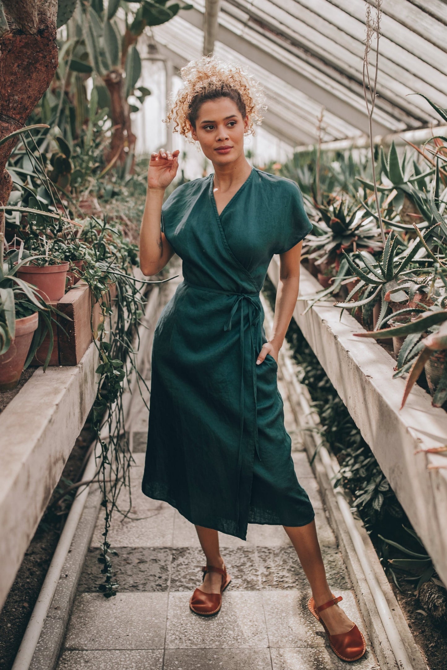 woman wearing green cap sleeve linen wrap dress with tie waist in greenhouse walkway