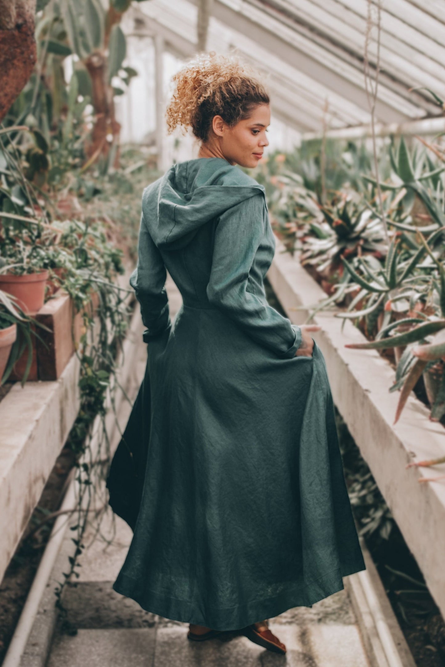 Woman wearing green heavyweight summer linen coat with hood and tie closure in greenhouse