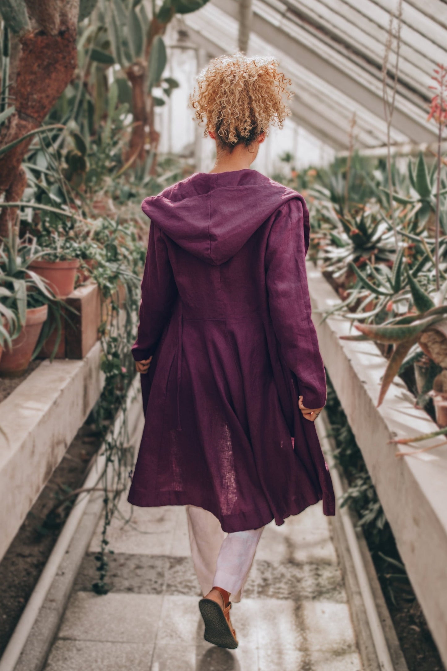 Woman wearing a purple midi length linen coat with hood and tie closure walking in greenhouse