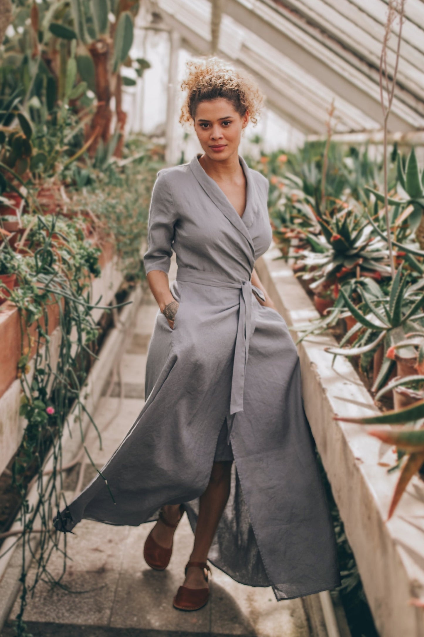 Woman wearing gray linen A-line dress with tie waist walking in greenhouse surrounded by plants