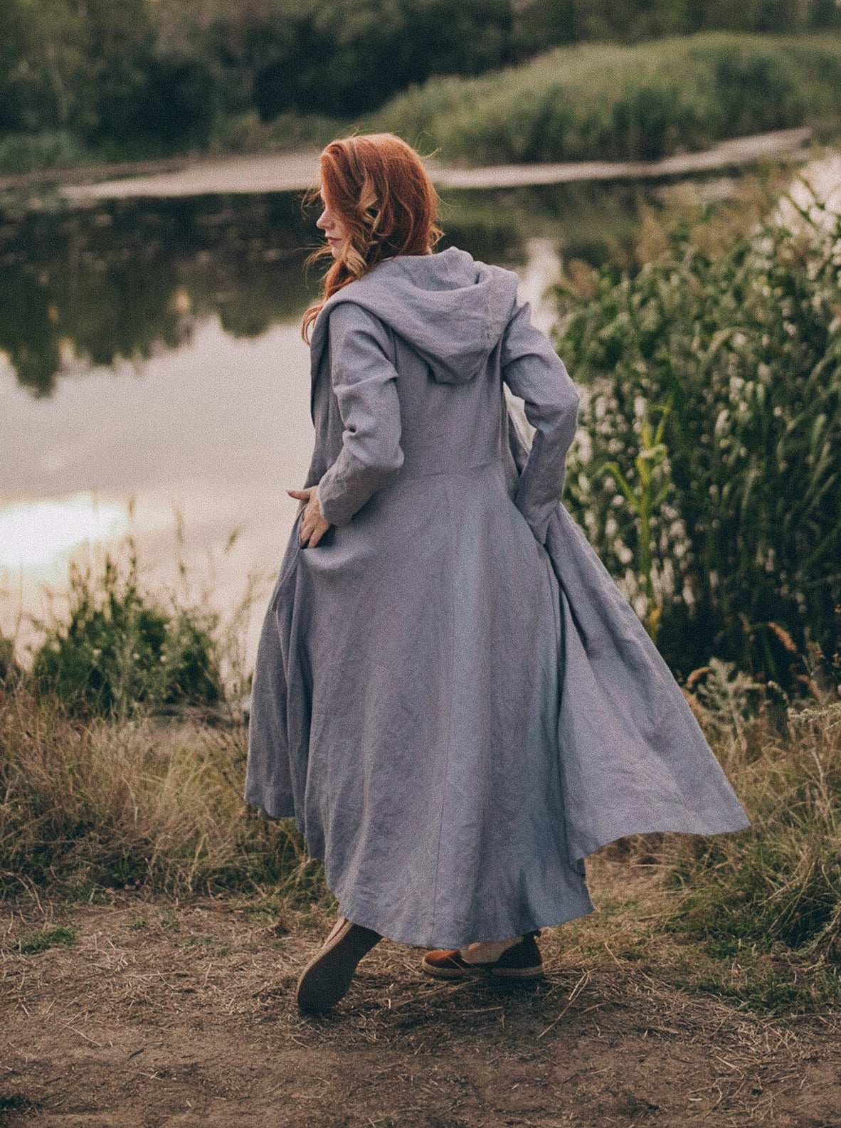 woman wearing heavyweight linen coat with hood standing near lake outdoors