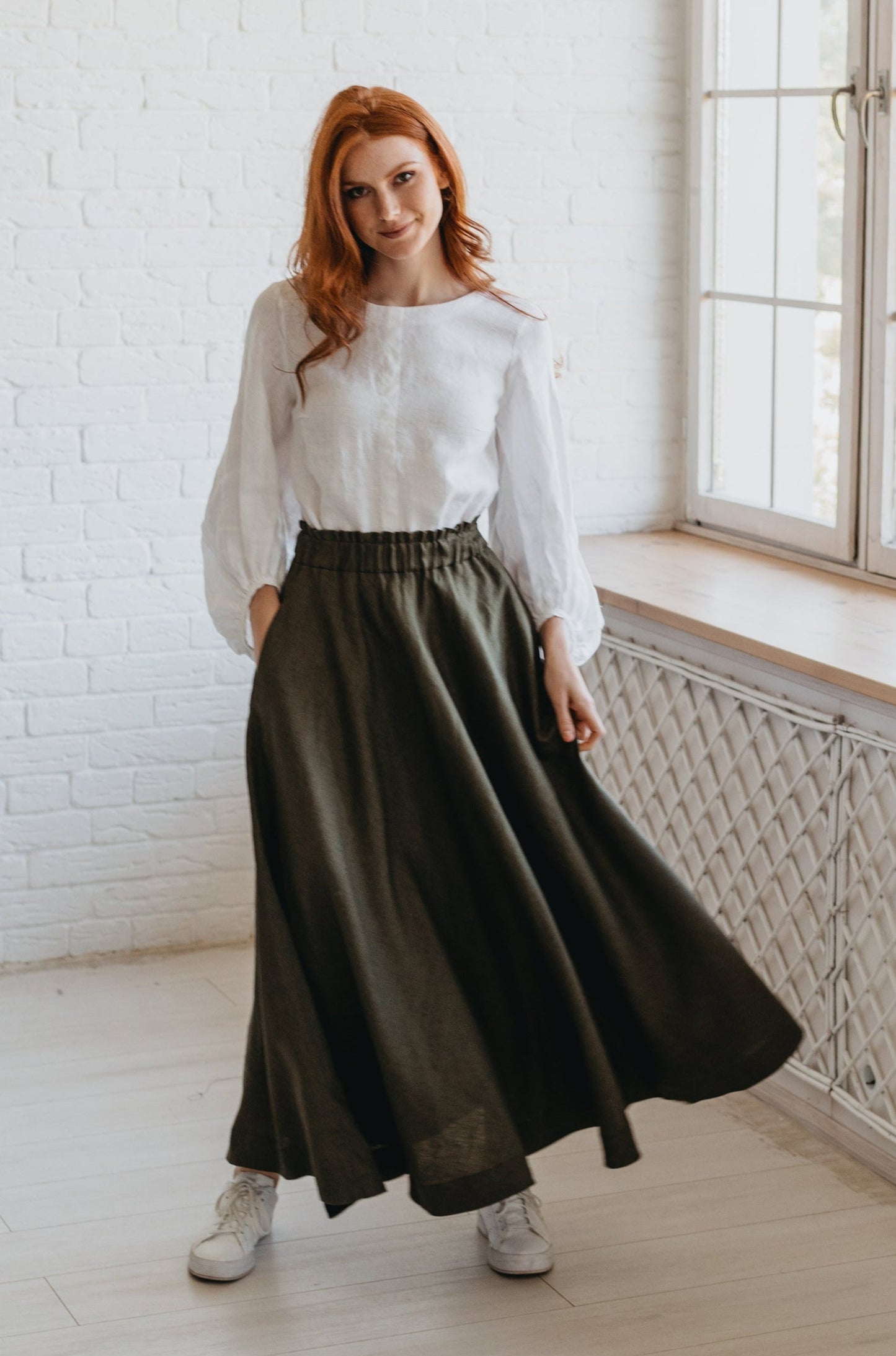 Woman wearing a heavyweight linen skirt with elastic waist and pockets, styled casually with a white blouse and sneakers indoors near window