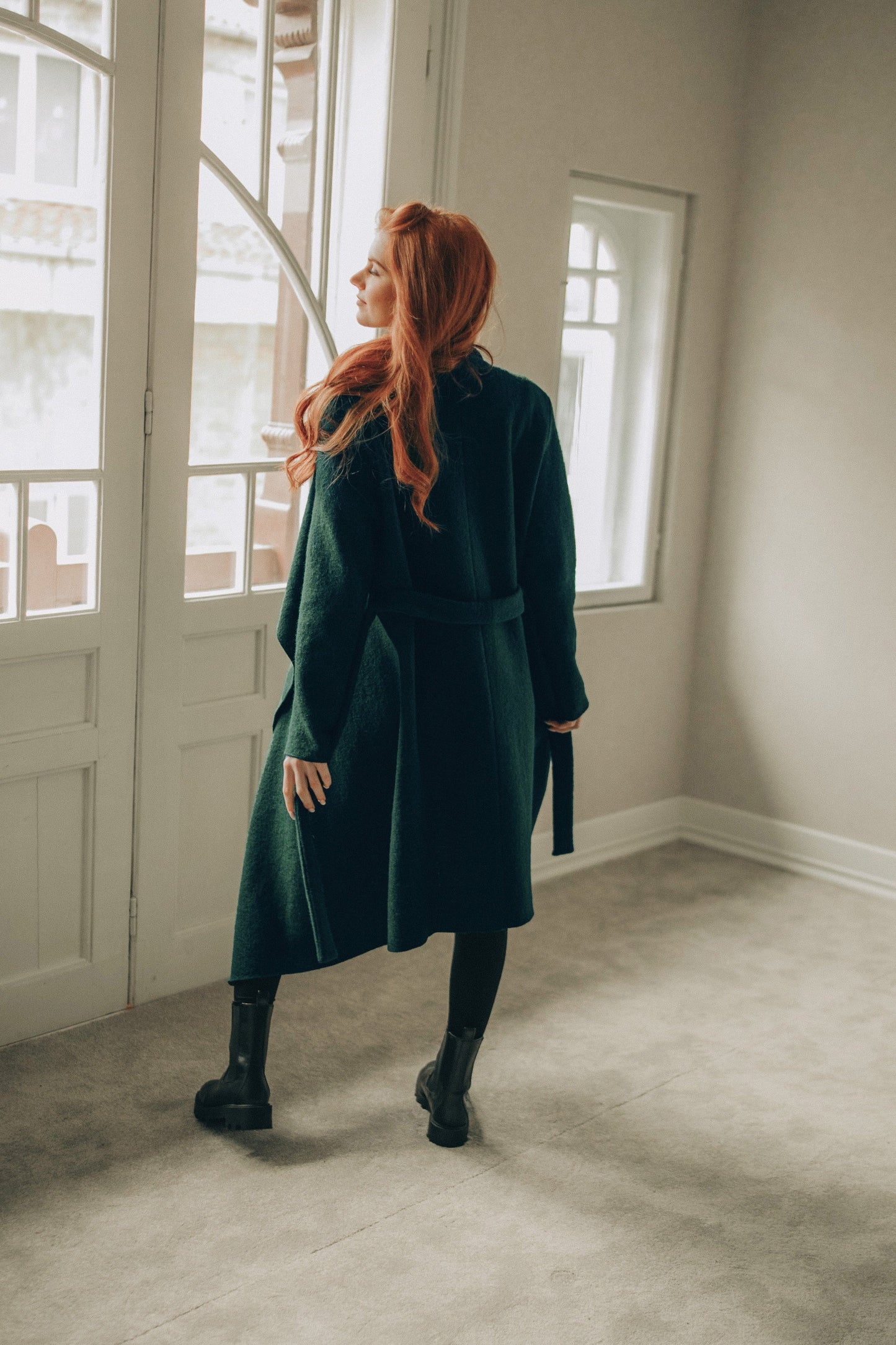 Woman with red hair wearing a green coat standing in a room with large windows.