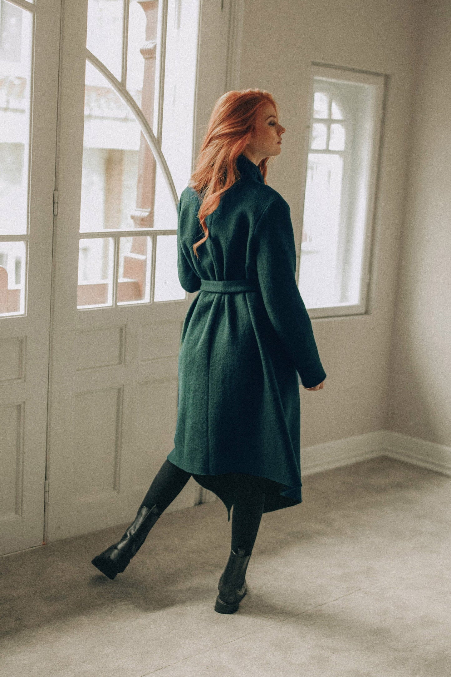 Woman in a green coat standing in a room with large windows.