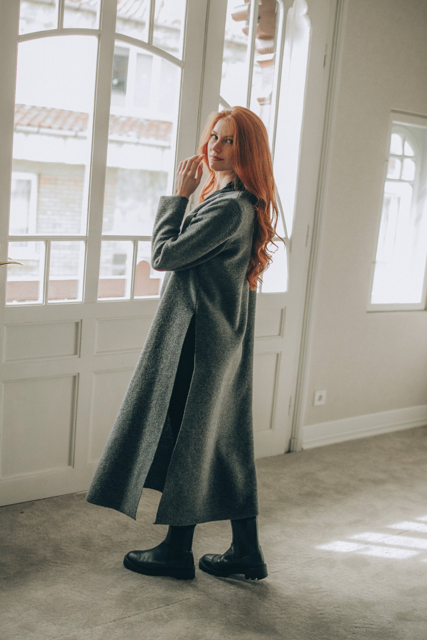 Woman in a long gray cardigan standing in a room with large windows.