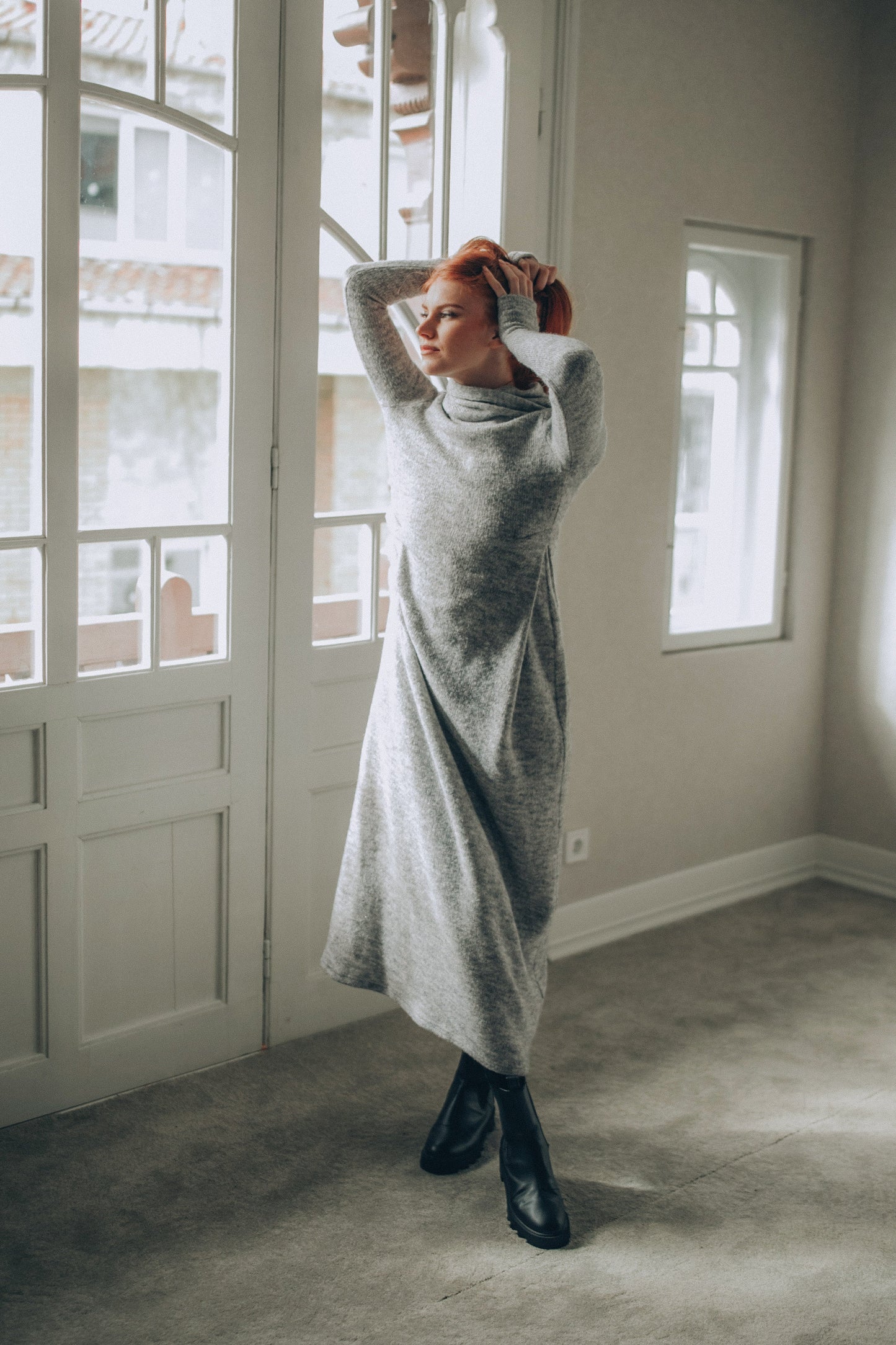 Woman in a long gray knitted dress standing in a room with large windows.