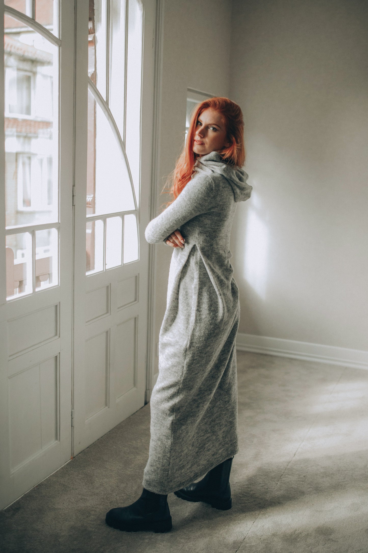 Woman in a long gray knitted dress standing in a room with large windows.