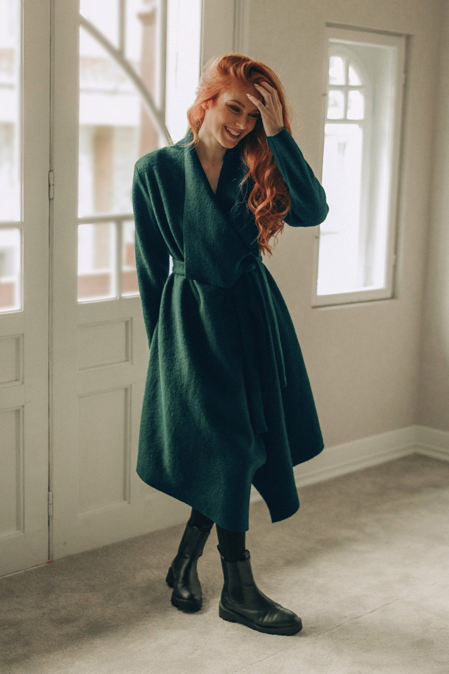 Woman in a green coat standing in a room with large windows.