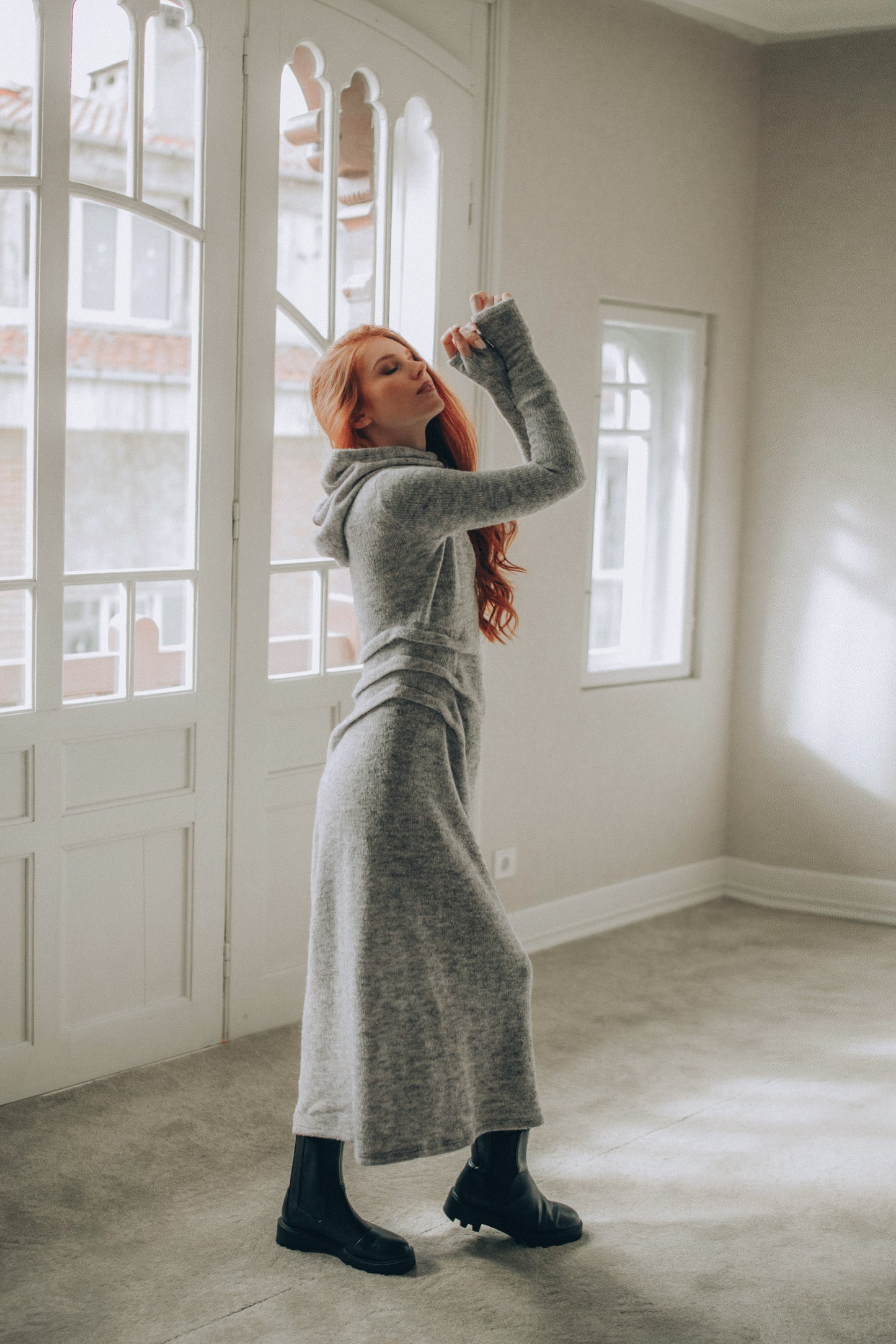 Woman in a long gray knitted dress standing in a room with large windows.