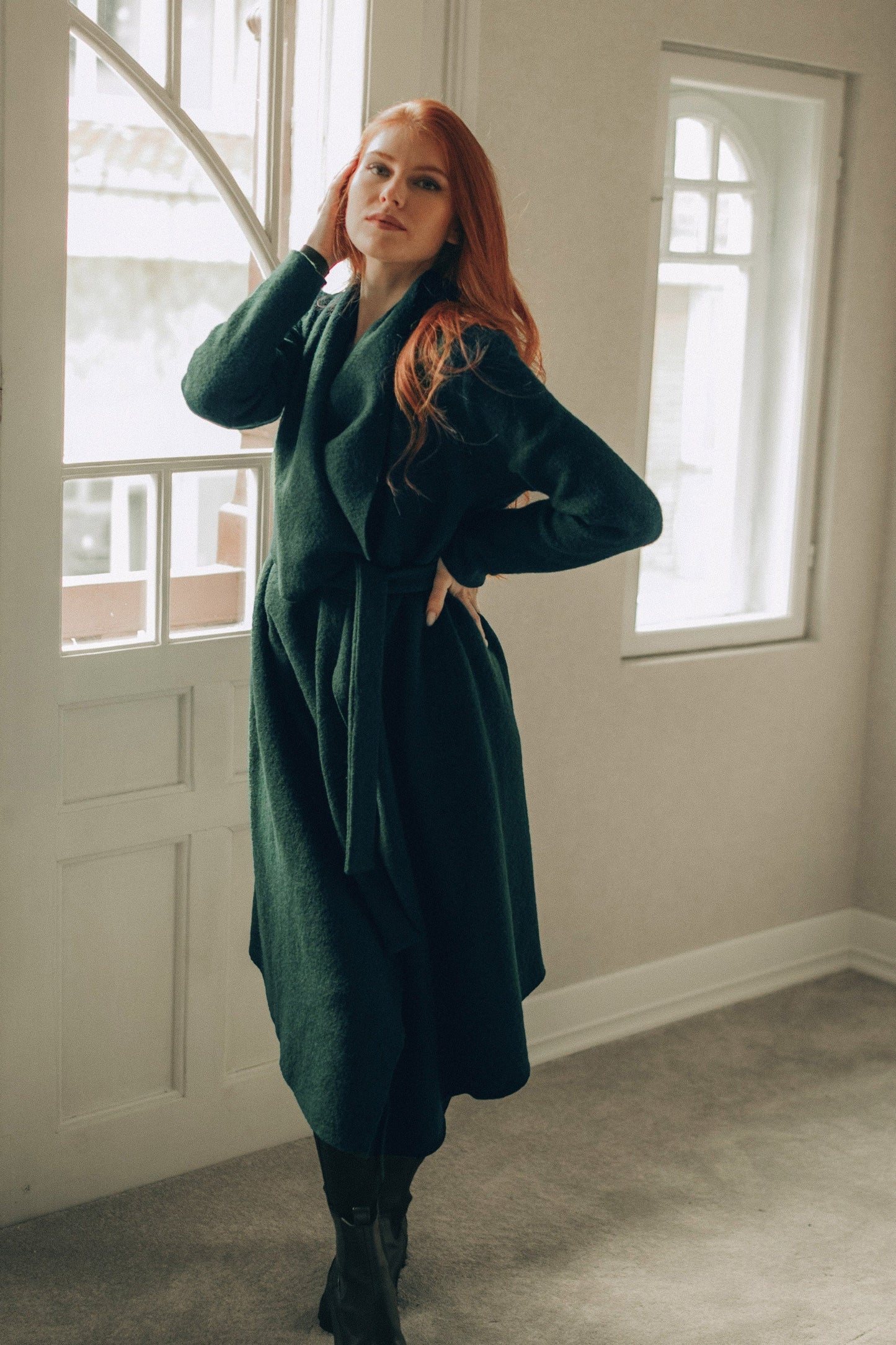 Person wearing a green coat standing in a room with large windows.