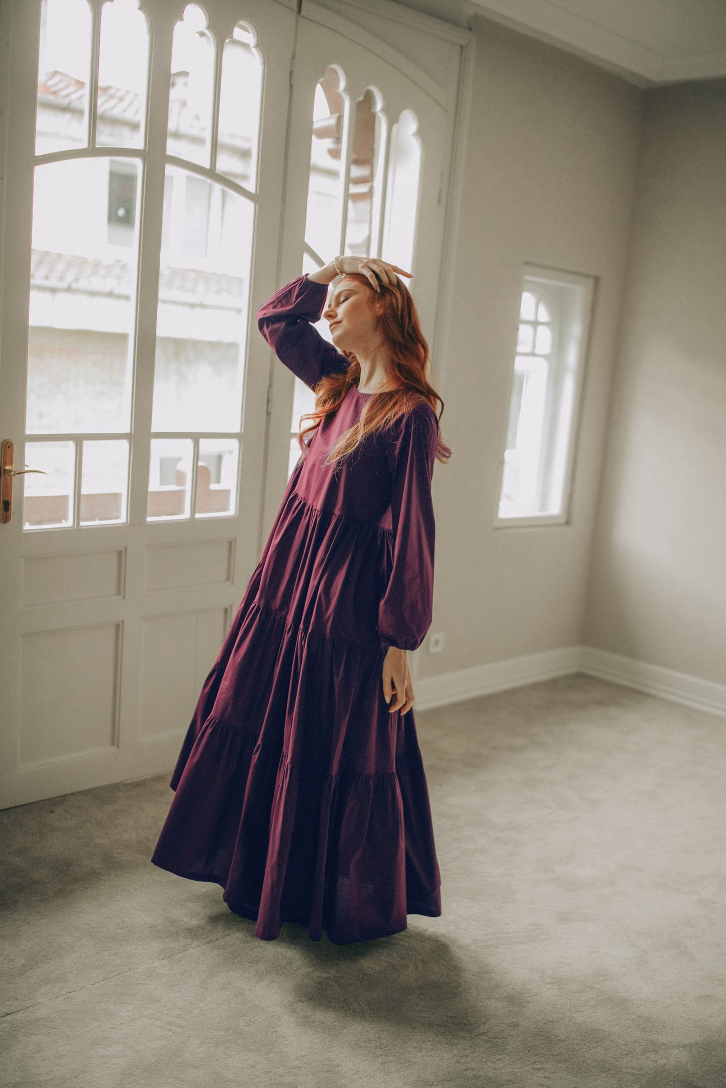 Woman in a long burgundy dress standing in a room with large windows.