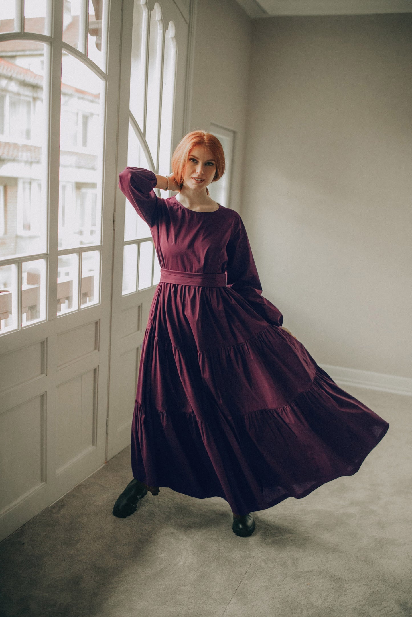 Woman in a long burgundy dress standing in a room with large windows.