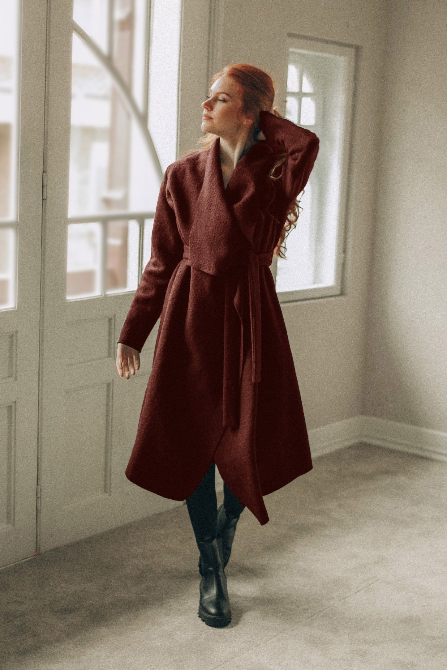Woman wearing a long brown cardigan coat in a room with large windows.