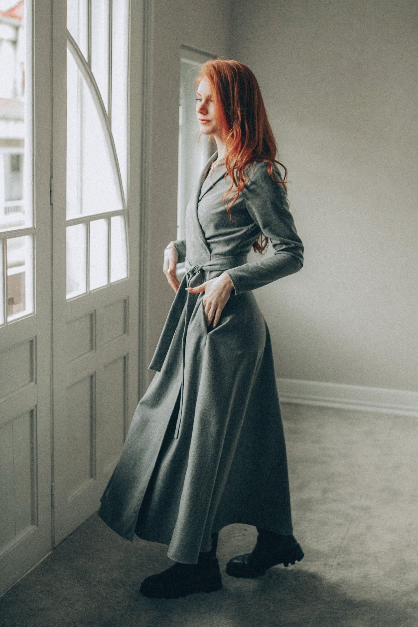 Woman wearing long gray wool wrap dress with shawl collar and side pockets indoors near window