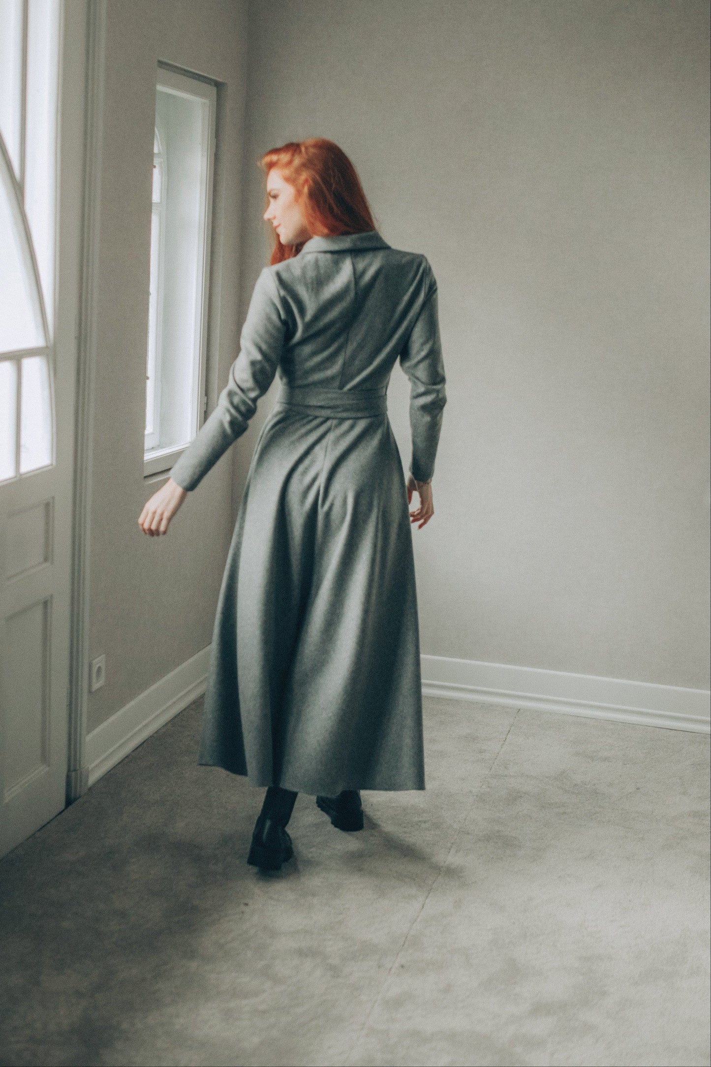 Woman in a long gray dress standing in a room with a window.