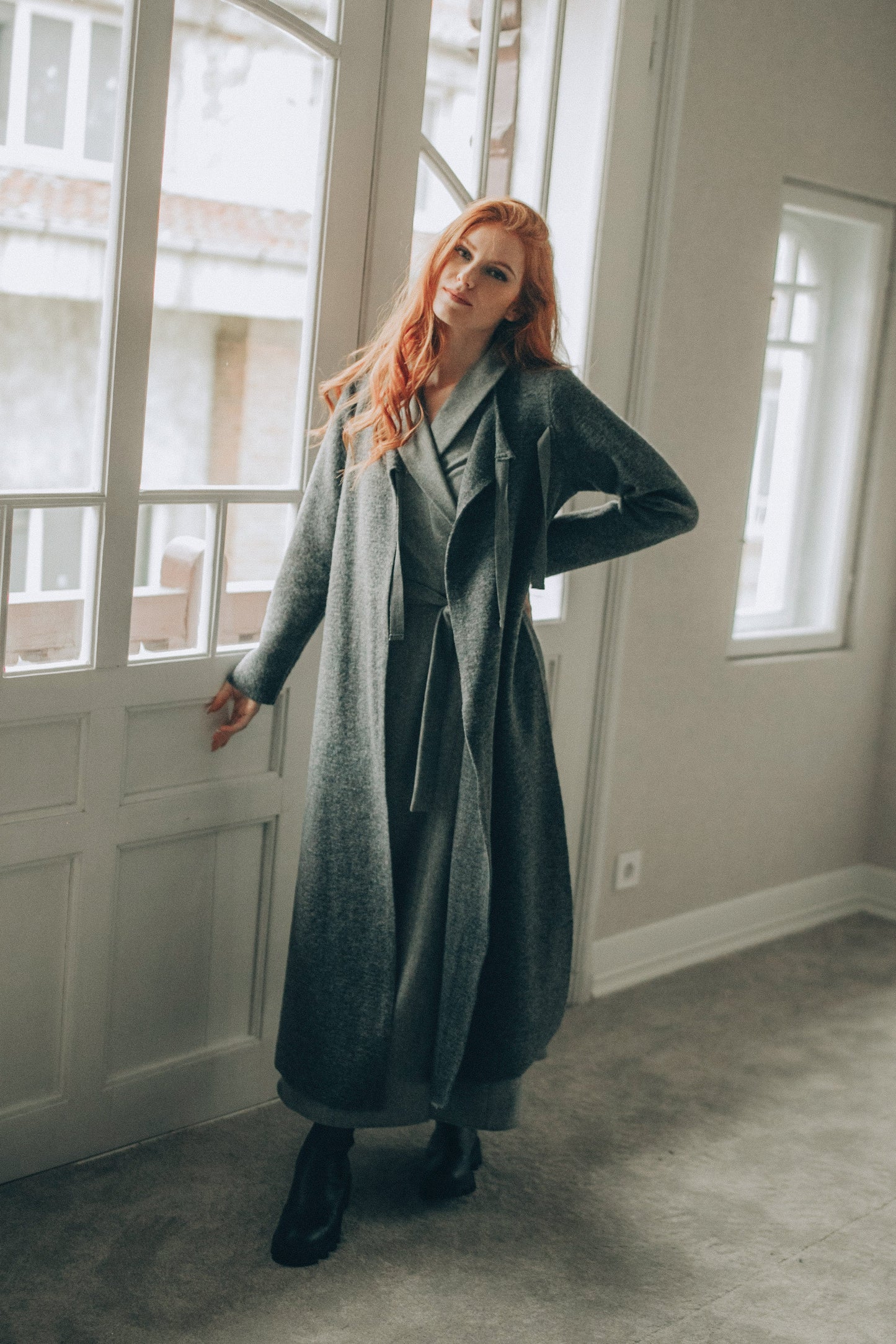 Woman in a long coat standing in a room with large windows.