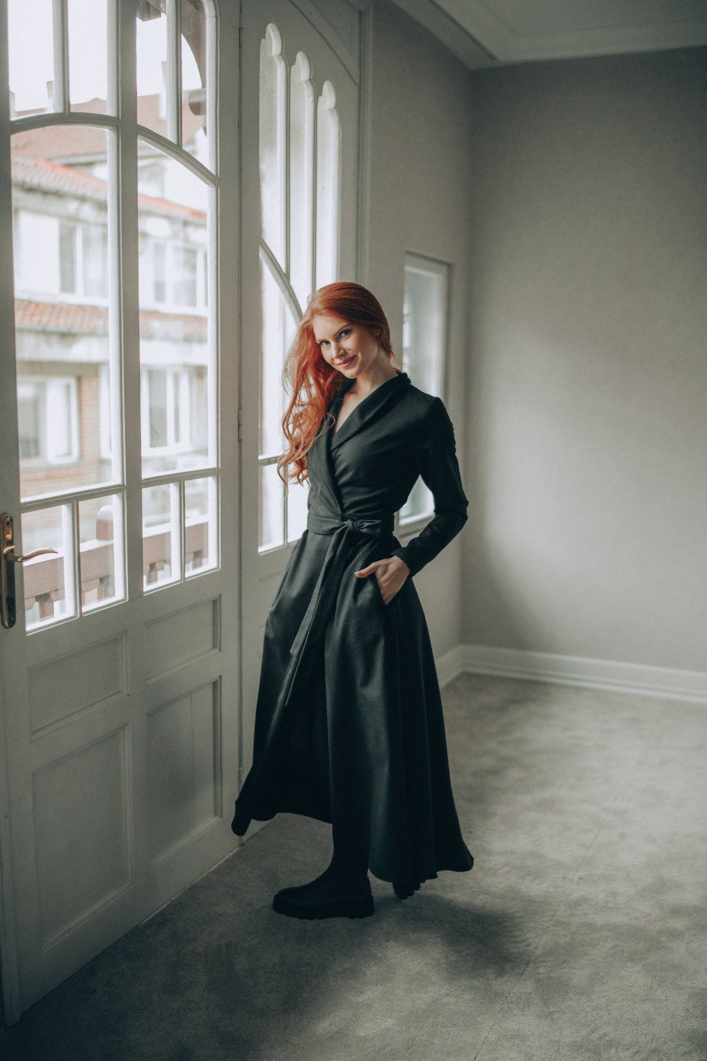 Woman in a wool wrap dress standing by a window in a room with light-colored walls.