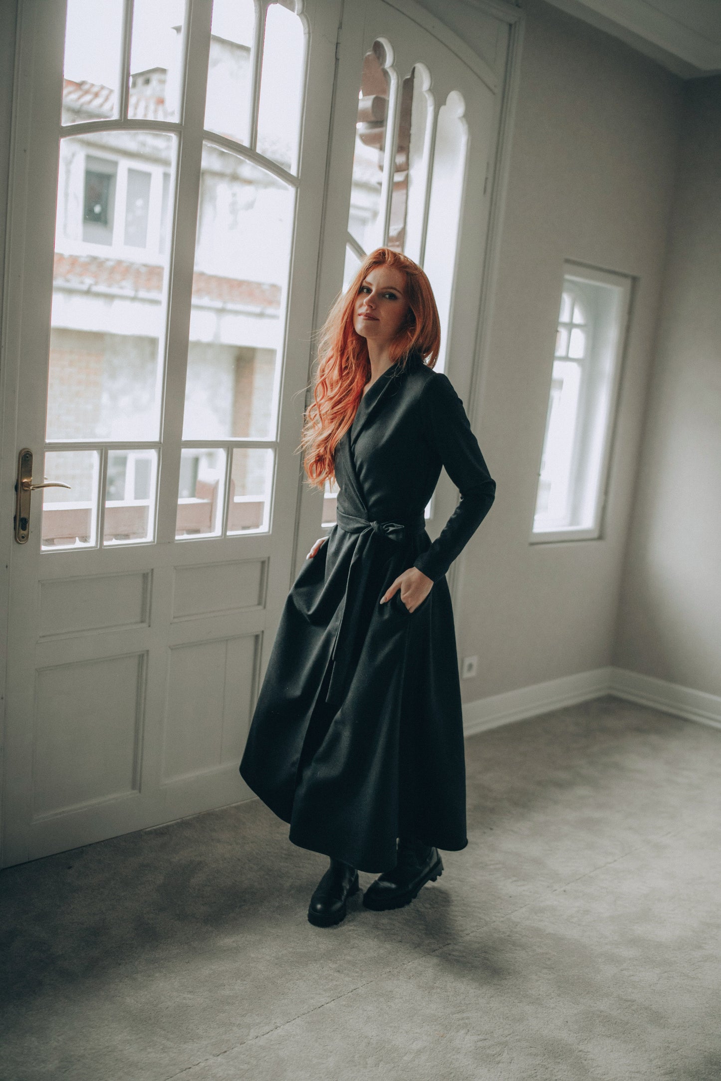Woman in a wool wrap dress standing by a window in a room with light-colored walls.
