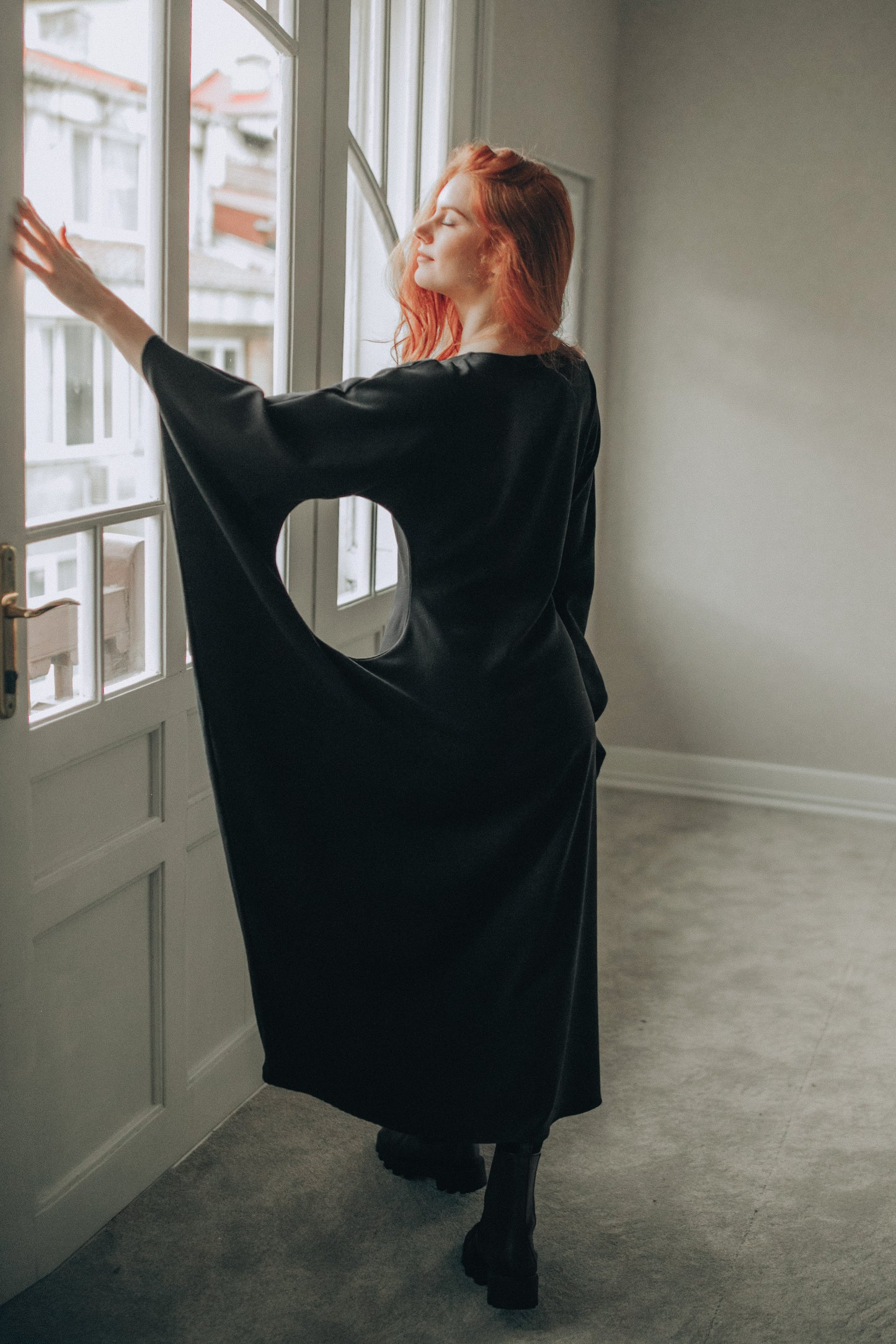 Person wearing a black dress standing by a window in a room with light-colored walls and carpet.