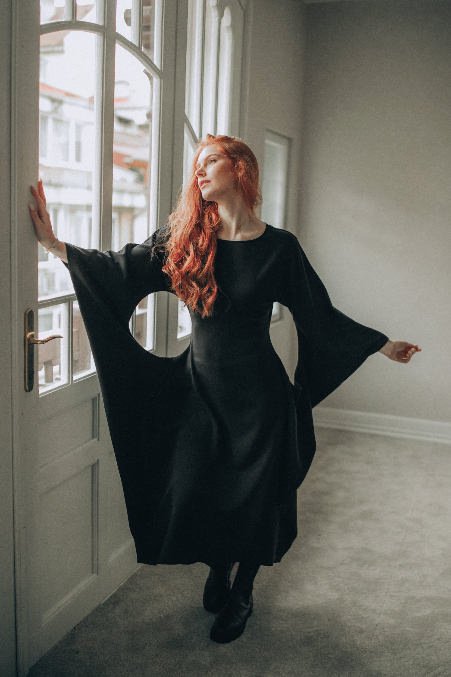 Woman in a black dress with wide sleeves standing in a room with large windows.