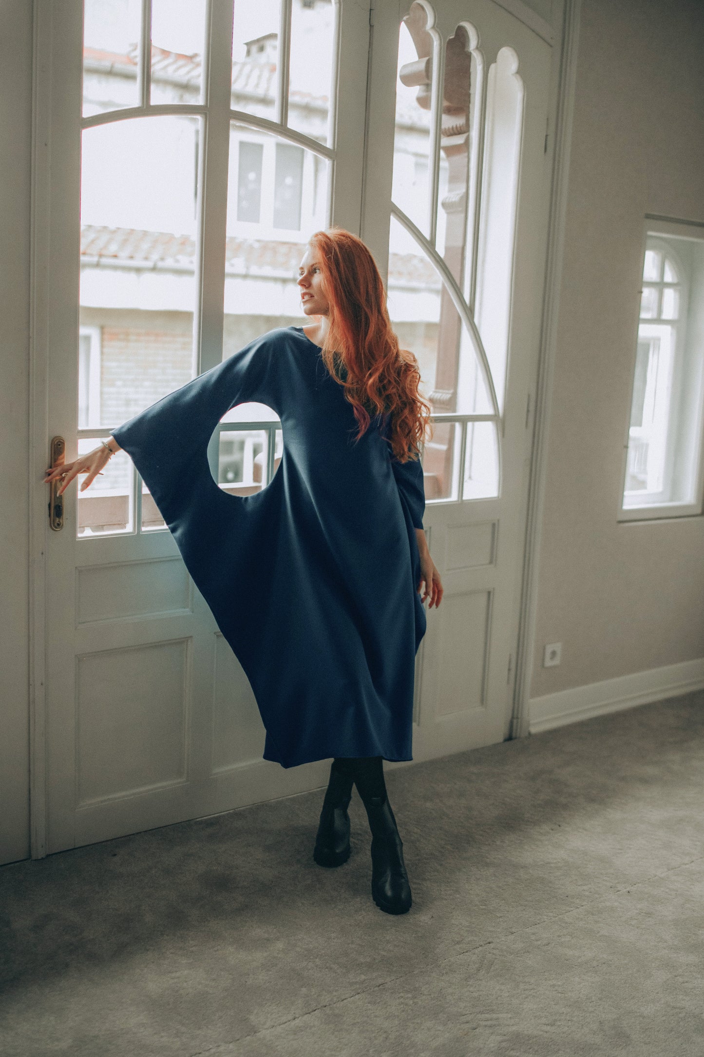Woman in a petrol blue dress standing in a room with large windows.