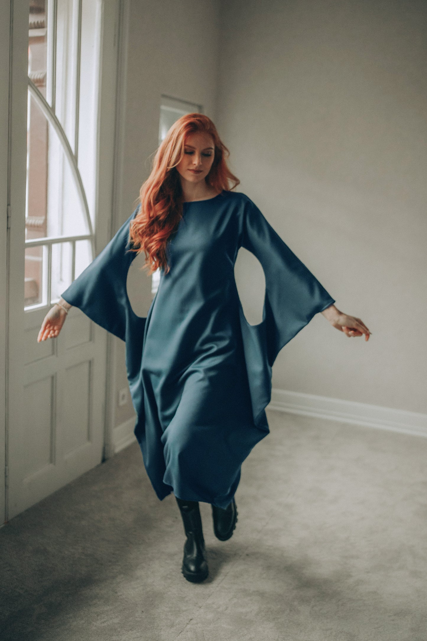 Woman in a petrol blue midi dress with wide sleeves standing in a room with a neutral color scheme.