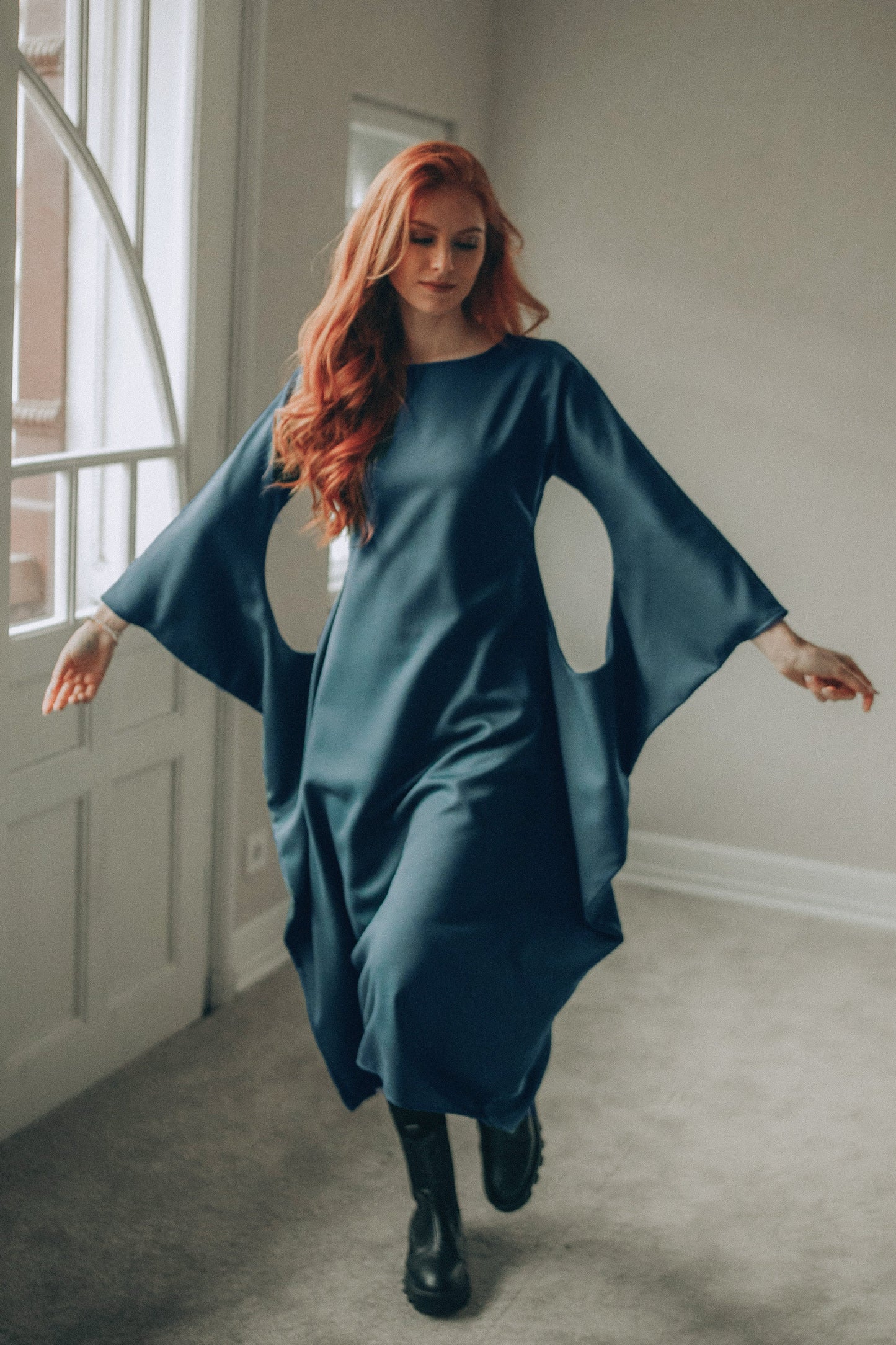 Woman in a petrol blue midi dress with wide sleeves standing in a room with a neutral color scheme.