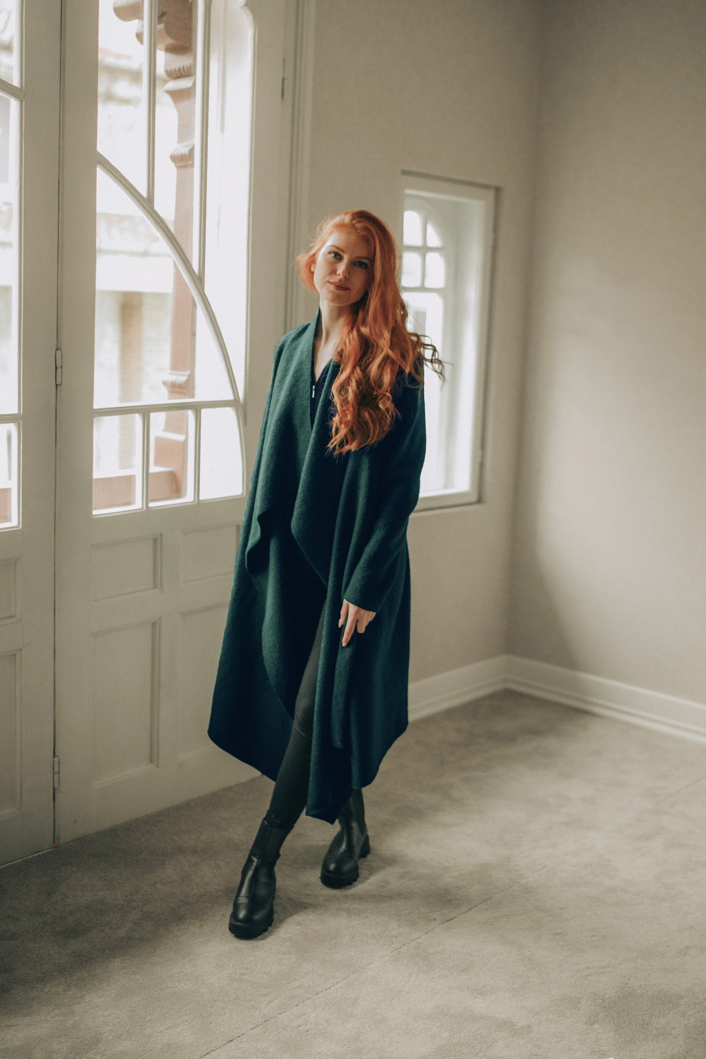 Woman in a green coat standing in a room with large windows.