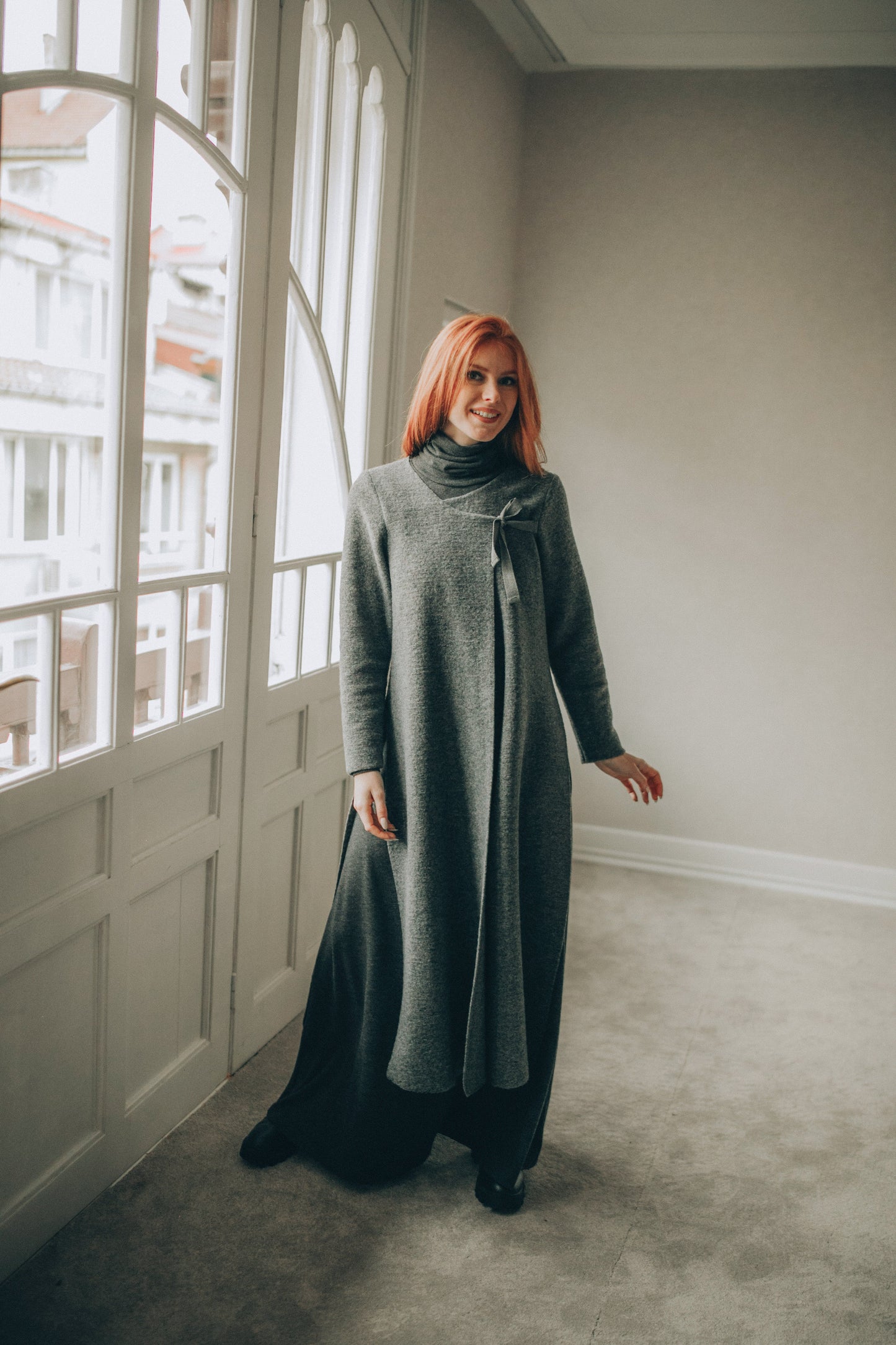 Woman in a long gray cardigan standing in a room with large windows.